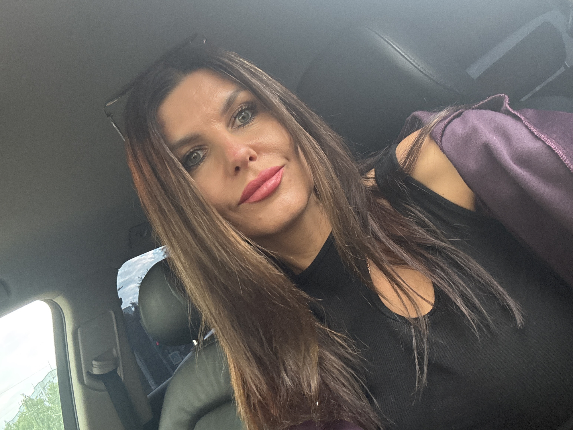 A woman with long, dark brown hair and green eyes takes a selfie inside a car. She is wearing a black top and a dark purple jacket draped over her shoulders. Her lips are painted a bright pink, and she is wearing makeup. She has glasses perched on top of her head. The interior of the car is visible, including a black leather seat and a seatbelt. Outside the car window, trees and a cloudy sky can be seen. The lighting suggests it is daytime.