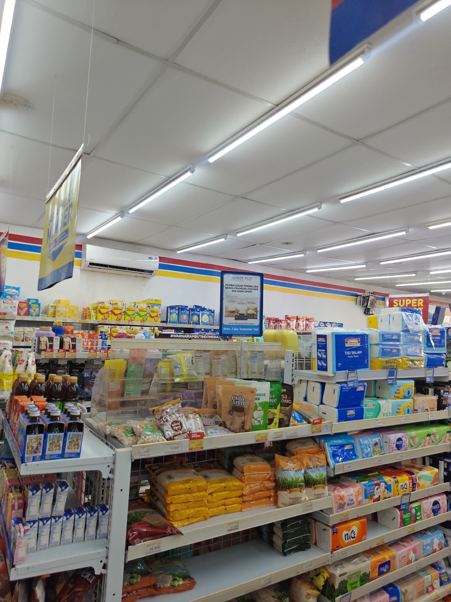 The image depicts the interior of a brightly lit convenience store, likely an Indomaret, given the branding and product selection. The store is stocked with a wide variety of goods, including packaged food items, beverages, toiletries, and household supplies, arranged on white metal shelves.
In the foreground, a refrigerated display case holds dark-colored bottled drinks. To the right, shelves are filled with packaged snacks, sugar, and facial tissues. Further back, rows of drinks in cartons are neatly stacked.
Prominently displayed are promotional signs and product information. A yellow sign hanging from the ceiling features text that is partially obscured. A blue informational poster is mounted on a wall in the background, detailing purchase limitations for specific rice products. The ceiling features a grid of white tiles and fluorescent lighting fixtures. An air conditioning unit is mounted on the wall above the shelves.
The scene is orderly and organized, suggesting a typical retail environment. The lighting is uniform and casts no significant shadows, indicating a controlled indoor setting. No individuals are visible in the image. The overall impression is one of a well-stocked and clean retail space.