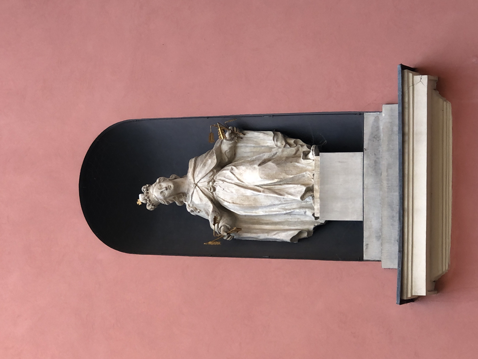 The image shows a stone statue of a crowned woman standing within an arched niche on a pink-colored wall. The statue depicts the woman wearing a draped gown and holding two golden arrows or scepters. She has a regal expression and her hair is styled in curls under her crown. The niche is dark, creating a contrast with the light-colored statue and the pink wall. Below the niche, there is a stone pedestal and a decorative ledge. This statue is located in Warsaw, Poland, specifically at the Church of St. Mary the Queen of Poland.