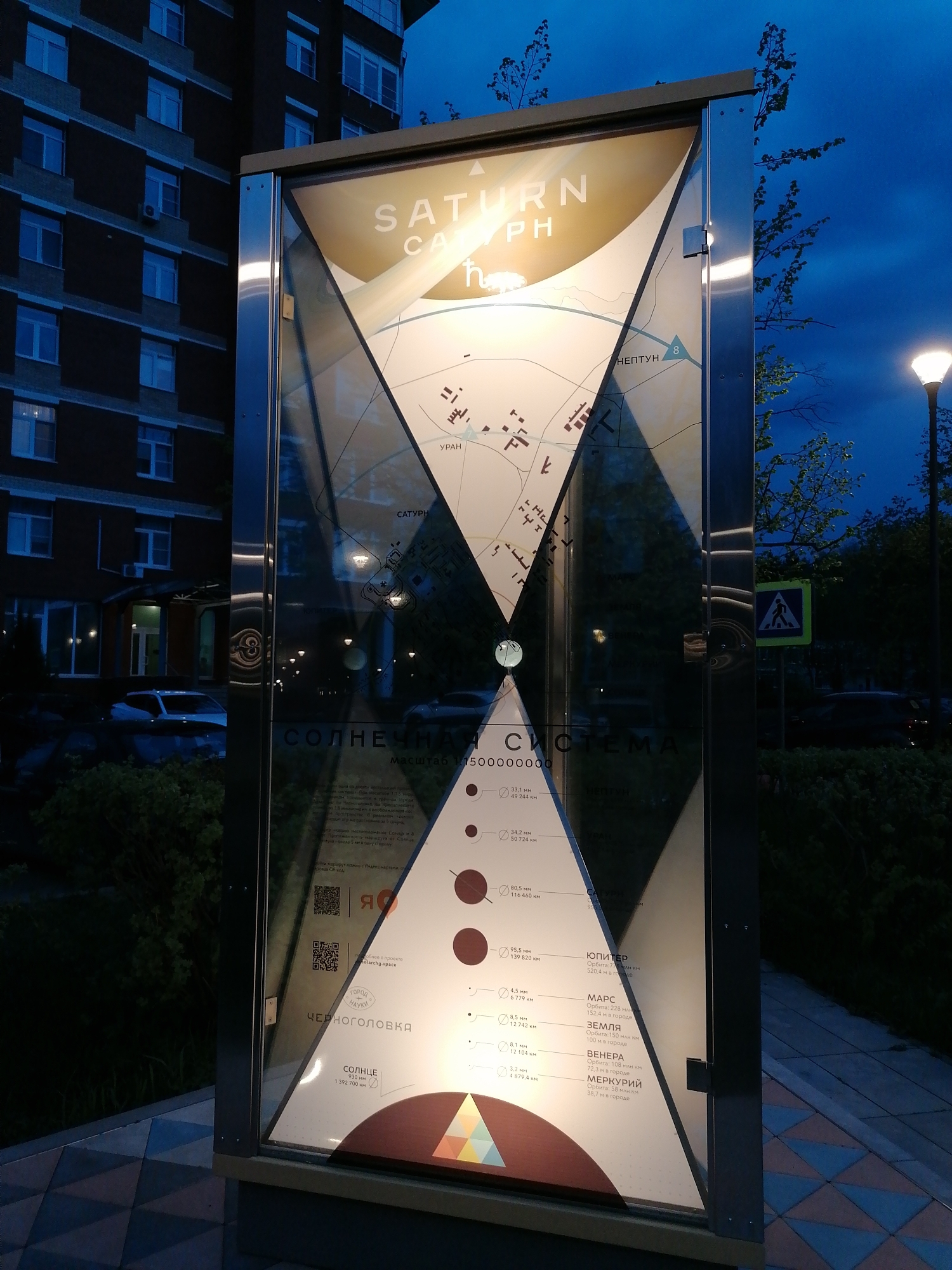 Here is a description of the image:
The image depicts an informational display related to the solar system, located in Chernogolovka. The display is constructed with a silver frame and features a graphic representation of the solar system, with information about the planets including Saturn, Uranus, Neptune, Jupiter, Mars, Earth, Venus, and Mercury. Each planet is represented by a circle and the sizes and distances are proportional in the scale of 1:5000000000. The top portion of the display also includes a map section. The text on the display is in Russian, identifying the planets. The sign also contains two QR codes near the middle.
The display is situated outdoors, with a building and street lamp visible in the background, as well as a pedestrian crossing sign. The sky is darkening. The informational display appears to be illuminated, adding brightness to the content presented on it.