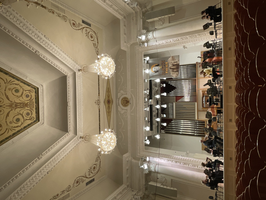 The image displays the interior of the Sverdlovsk State Academic Philharmonic Hall in Yekaterinburg, Russia, viewed from an audience perspective facing the stage. The hall features an ornate, light-colored ceiling with decorative moldings, stucco work, and painted floral patterns in gold and reddish-brown hues. Two large, illuminated crystal chandeliers hang centrally from the ceiling, flanked by additional decorative motifs.
On the elevated stage, an ensemble of approximately twelve musicians is performing. Instruments visible include multiple violins/violas, a cello positioned on the far left, a double bass on the right, and a yellowish wind instrument (possibly a saxophone or oboe) to the right of the central performers. A piano or harpsichord is situated towards the rear-center of the ensemble. Most musicians wear dark, formal clothing, while one individual on the right side of the stage is dressed in a yellow top. All musicians are seated, actively playing their instruments, and observing sheet music on individual stands. The stage is well-lit by multiple overhead spotlights.
Behind the musicians, a large pipe organ serves as the backdrop, its numerous vertical pipes varying in color from silver to reddish-brown. A vertical banner featuring a portrait of Johann Sebastian Bach and the text "BACH FEST" hangs to the right of the organ pipes. The foreground of the image shows several rows of unoccupied, dark red or brown upholstered audience seats.