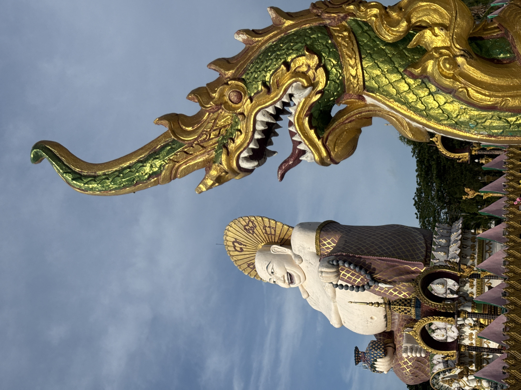 In Baan Plai Laem, Thailand, a large, smiling Buddha statue sits to the left of the frame, adorned with a golden halo and a string of dark beads. To the right, a vibrant green and gold serpent statue with sharp teeth and a forked tongue looms large, its head angled towards the Buddha. The statues stand against a backdrop of a bright blue sky with wispy clouds and a line of green trees in the distance. The foreground is partially obscured by decorative golden fencing with pink triangular flags.