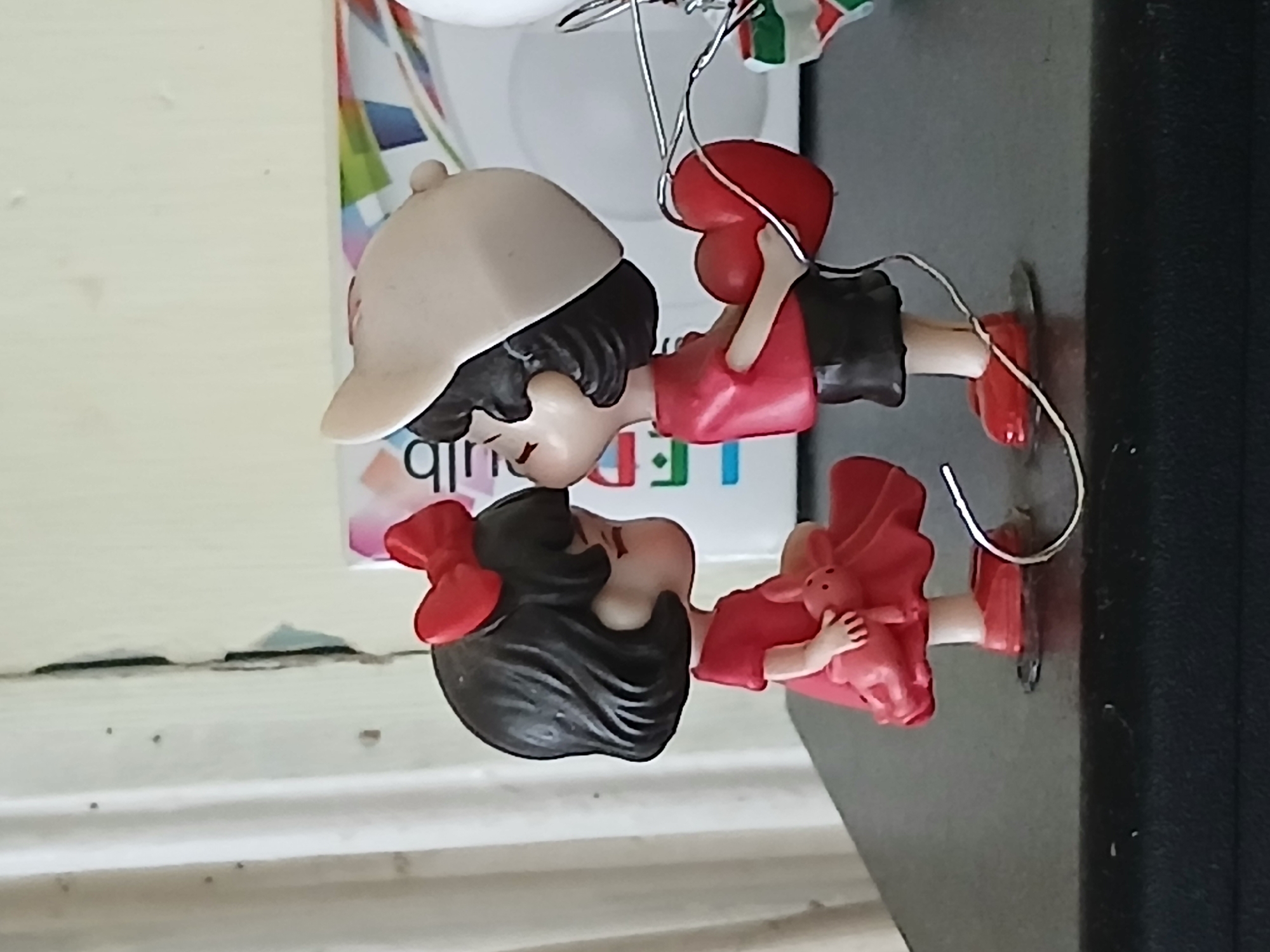 A close-up, medium shot, taken at an eye-level perspective, shows two small figurines on a dark, flat surface, likely a shelf or table. The figurines are posed in a romantic embrace, with their faces close together as if about to kiss. The figurine on the left is a girl with short, dark hair adorned with a red bow. She wears a red dress and holds a small pink bunny. The figurine on the right is a boy wearing a red shirt, black shorts, and a light-colored baseball cap. He holds a red heart. The background consists of a textured, light-colored wall with vertical lines and a partially visible cardboard box with colorful abstract shapes and lettering. The lighting appears to be from the front, casting soft shadows. There are no visible elements that indicate the image was taken in Telavi, Georgia.