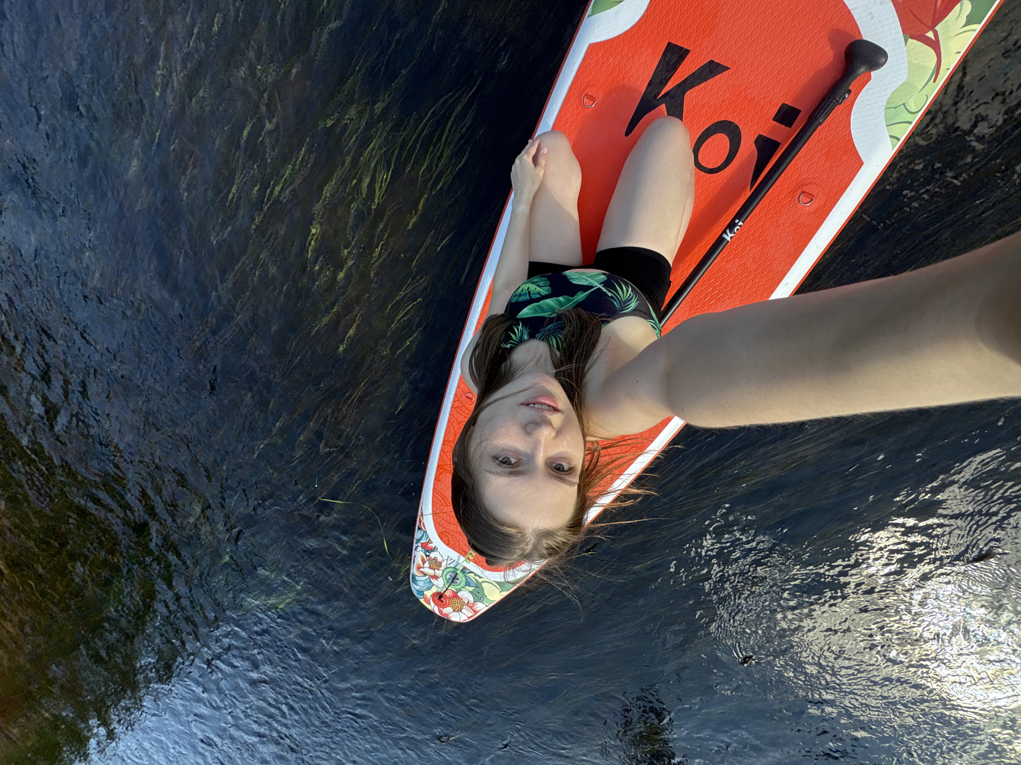 A young woman with long brown hair is paddleboarding on a river. She is wearing a black swimsuit with a green leaf pattern and black shorts. She is holding a paddleboard with an orange and white design. The water is dark and appears to have aquatic vegetation. The scene appears to be a natural outdoor setting. The location is Siversky, Russia.