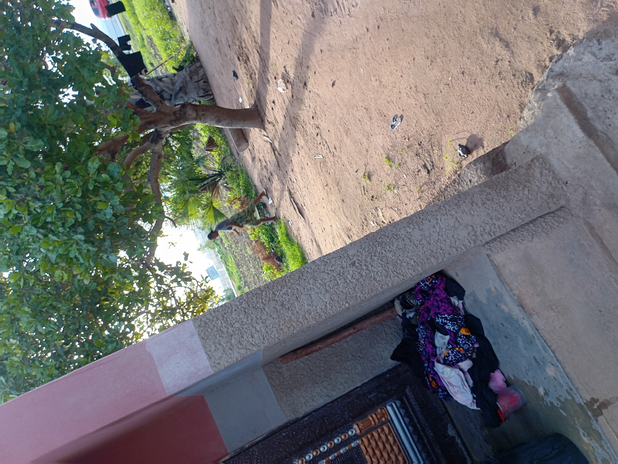 The image shows an outdoor scene with a building in the foreground and a tree and vegetation in the background. A person wearing a patterned skirt and a t-shirt is walking away from the camera in the middle ground. Clothes are hanging on the branches of the tree. In the foreground, a pile of colorful clothes is on the ground next to a red bucket. The ground appears to be dirt or sand. In the far background, a red car is visible. The lighting suggests it is daytime, with sunlight casting shadows.