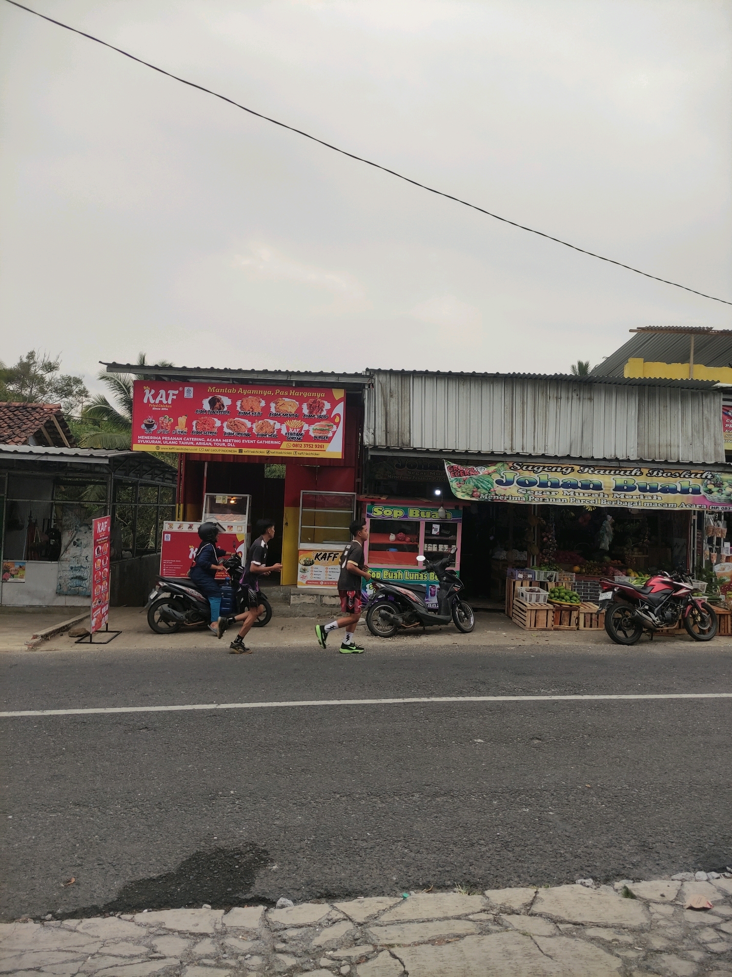 In Wadaslintang, Indonesia, a street scene unfolds with a row of businesses lining the side of a paved road. The businesses include a food stall with a prominent "KAF" sign advertising fried chicken and a fruit stand displaying various produce. A few motorcycles are parked in front of the establishments, and two individuals are captured in motion, one riding a scooter with a passenger, and another person running on the sidewalk. The sky above is overcast, contributing to the overall muted lighting of the scene. The environment appears to be a typical commercial area, with buildings and street infrastructure visible.