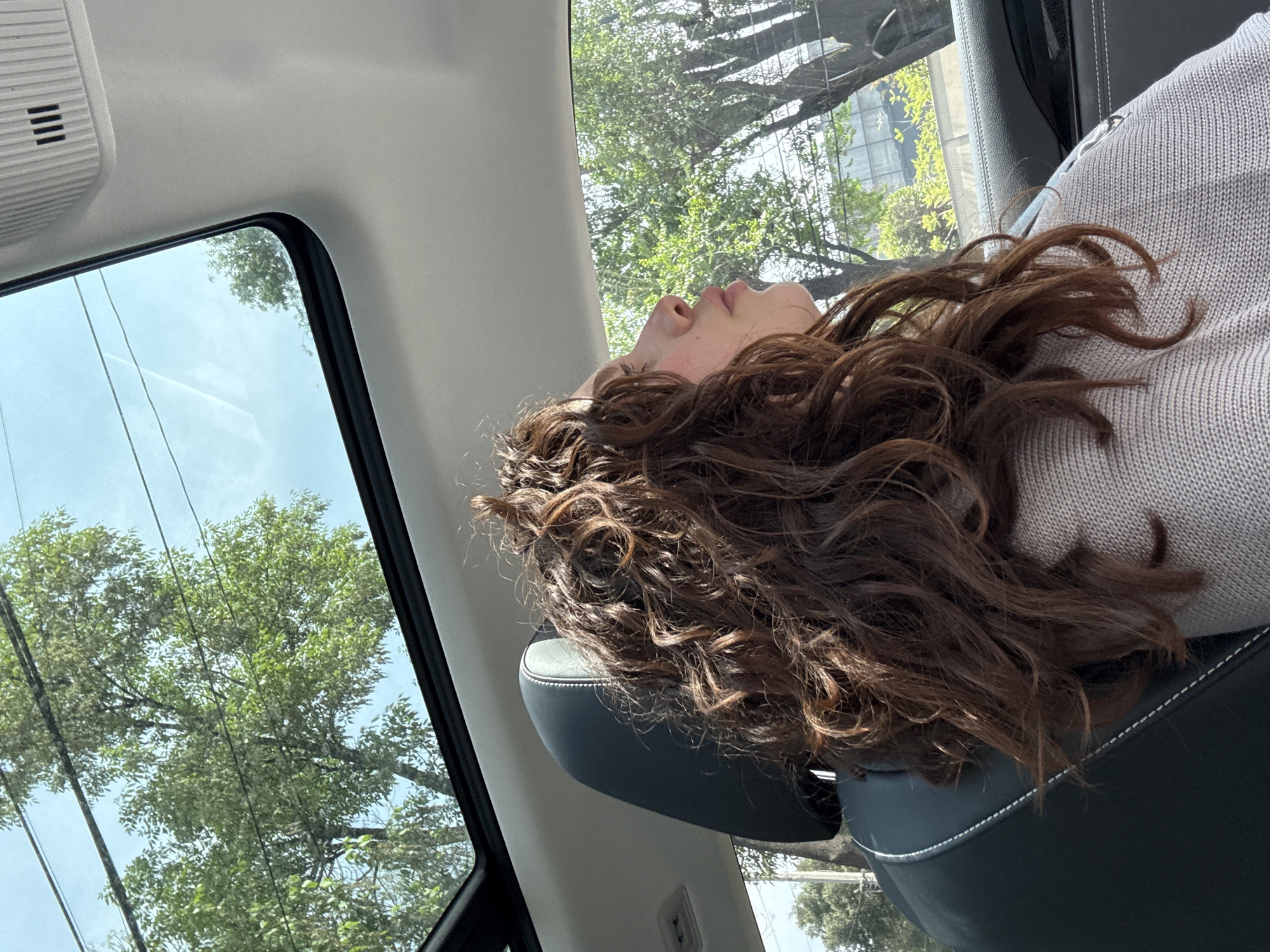 A woman with long, curly, brown hair is reclining in the passenger seat of a vehicle, presumably a car. She is positioned against the headrest. A grey sweater is visible. Through the window, trees are visible and buildings which, based on architecture and vegetation are typical to Mexico City. Overhead electrical wires are also present. The interior of the car includes the trim and seating upholstered in dark leather. A light-colored interior panel with ventilation slats is near the top left of the frame.
