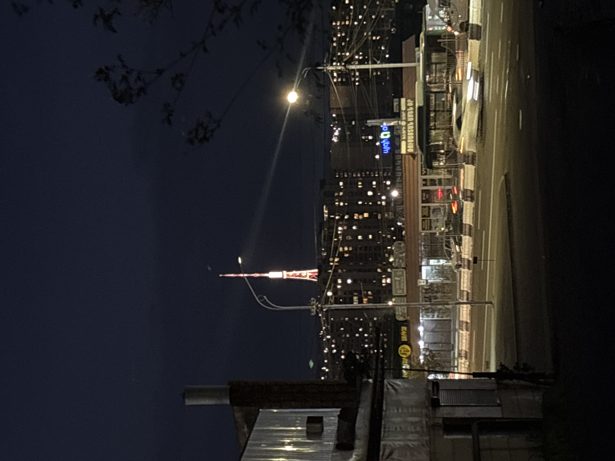This media file captures a nighttime urban scene in Yerevan, Armenia. The image, correctly oriented, displays a city street at dusk or night, illuminated by artificial lights.
In the mid-ground, slightly to the left of the center, stands the prominent red and white Yerevan TV Tower, a distinct landmark, brightly lit against the dark sky. Below and to its right, multi-story buildings line a street, their windows glowing with warm light, suggesting active residences or businesses.
The foreground on the left features dark, silhouetted tree branches and leaves against the deep blue-black sky. On the right side, a busy street is visible, with traffic indicated by blurred lights of vehicles. A bus is partially discernable on the road. The road surface appears wet, reflecting the bright white headlights and taillights of passing cars, as well as the overhead streetlights. A central barrier or median with black and white stripes separates lanes.
Numerous streetlights illuminate the scene, with one particularly bright lamp in the mid-left casting a visible flare. Overhead wires and cables crisscross between utility poles and buildings. In the immediate foreground, indistinct parts of rooftops or industrial-looking structures are visible.
Several signs with Armenian text are visible on the building facades. A prominent blue sign reads "ՄԱՅՐ ՈՐԱԿ" (Mayr Vorak, meaning "Mother Quality" or "Main Quality") and "ՀԱՂԹԱՆԱԿԻ ՀՐԱՊԱՐԱԿ" (Haghtanaki Hraparak, meaning "Victory Square"), clearly indicating the location as Yerevan. Another smaller sign below it appears to say "ՎԵՐՋԻՆ" (Verjin, meaning "Last"). A circular yellow sign with Armenian text is also partially visible on a building further down the street. The scene depicts typical urban activity after dark, with illuminated structures and moving traffic.
