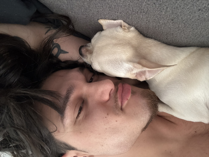 The image shows a young adult male and a small dog in close proximity, lying on a dark gray, textured surface, likely indoors. The male, positioned on his side, has long, dark, wavy hair and stubble on his chin and jawline. His left eye is partially open, directed towards the viewer, and his lips are slightly parted. A dark, stylized tattoo is visible on his left upper arm. Resting against the man's lower face and neck area is a short-haired, light-colored dog, consistent with a Chihuahua. The dog's head is facing away from the viewer, and its ears are erect. The scene depicts a moment of repose or close companionship. No visual information within the image allows for the identification of a specific location in Sofia, Bulgaria.