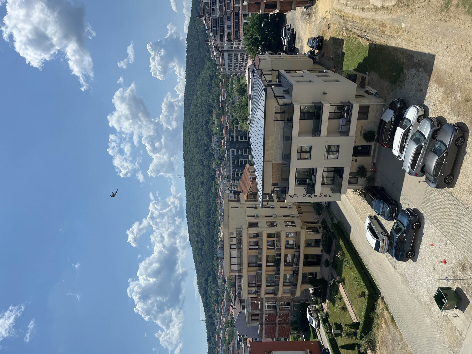 This is a high-angle, full shot of a residential area in Obzor, Bulgaria, on a sunny day. The scene is dominated by a densely forested hill that rises behind a cluster of buildings. Several multi-story apartment buildings with modern architecture and balconies are visible, interspersed with smaller houses with red-tiled roofs. A paved road runs alongside one of the apartment buildings, with several cars parked on it. The sky is a vibrant blue, with fluffy white clouds scattered across it. A single bird is captured in flight against the clouds. The sun casts long shadows, indicating it is likely late afternoon. The overall impression is one of a peaceful, developed hillside community under a clear, bright sky. A partially visible sign on one of the buildings reads "PARADISE".