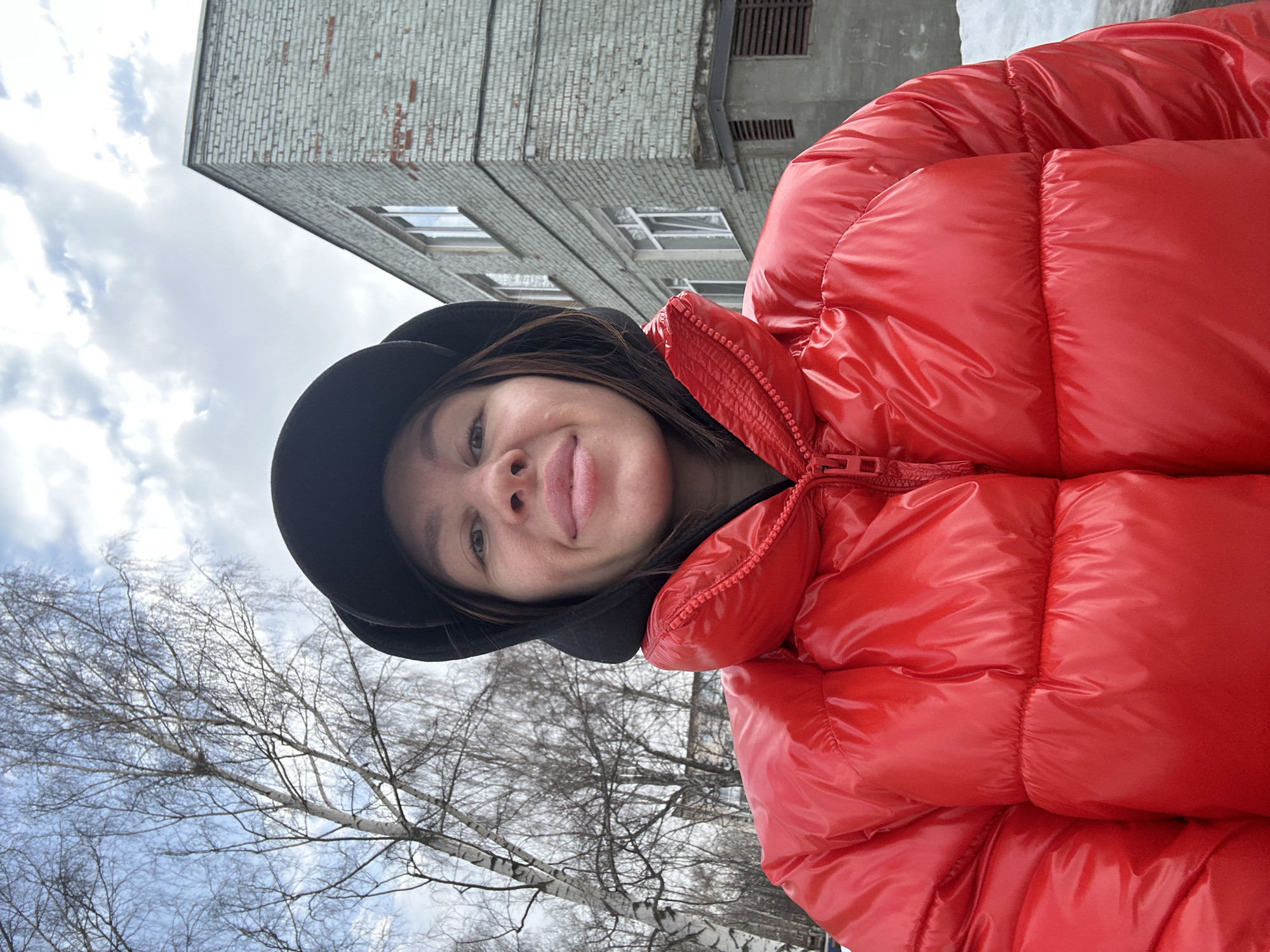 An adult female is depicted in a close-up selfie, facing the camera directly with a neutral expression and slightly parted lips. She wears a shiny, bright red puffer jacket, fully zipped, and a black baseball cap over a black hoodie, with visible brown hair. The scene is outdoors, in the city of Votkinsk, Russia. The background features a partially cloudy sky. To the subject's left, bare deciduous trees with light-colored bark stand, indicating a cold season. To her right, a multi-story building constructed of light grey brick with white-framed windows is visible, along with a utility structure featuring a louvered vent at its base. Patches of snow are present on the ground to the right, adjacent to the building.