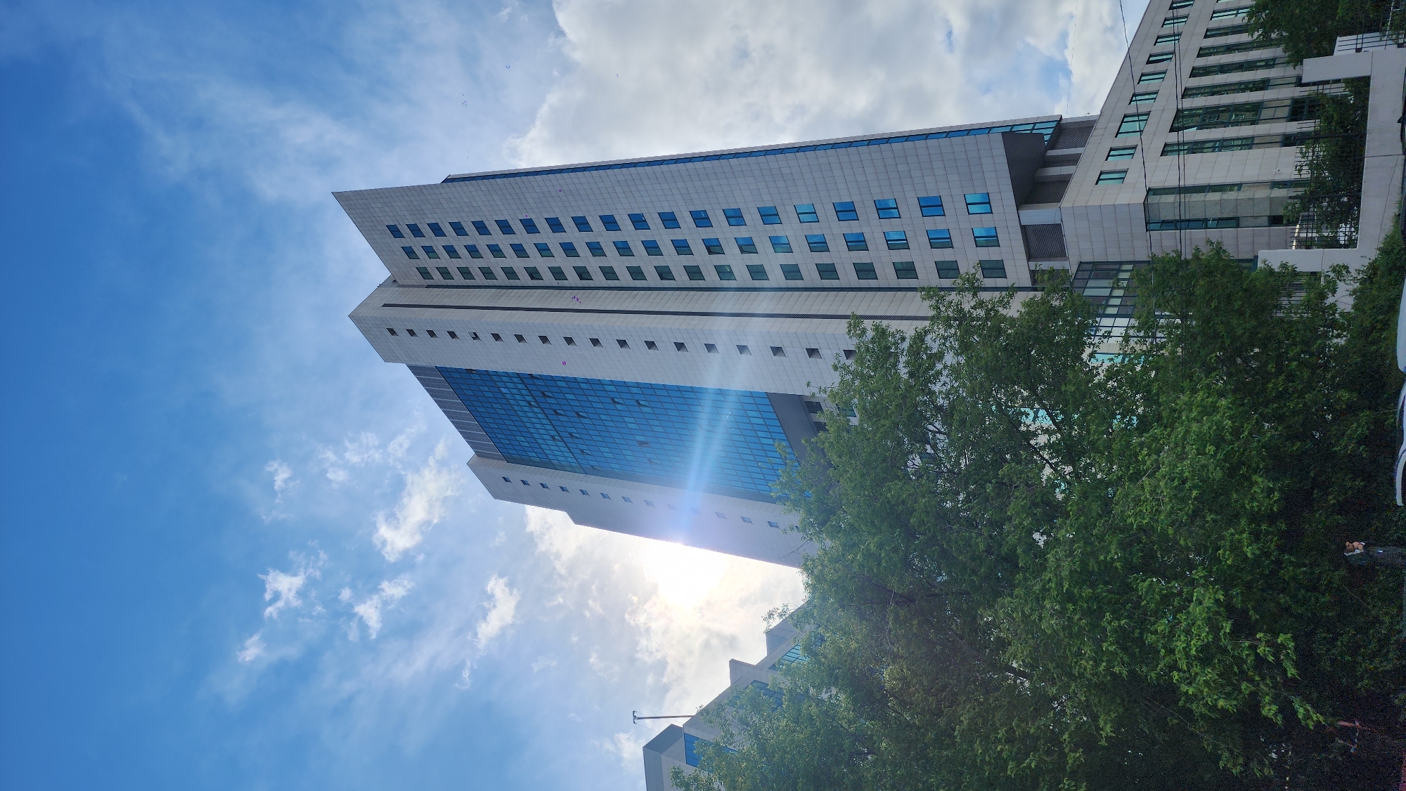 A low-angle shot captures a modern building in Moscow, Russia, under a sky with scattered clouds. The building features a light-colored exterior with blue-tinted windows. The sun shines behind the building, creating a lens flare effect. Green trees partially obscure the view of the building's lower section. The overall composition emphasizes the building's height and contemporary design.