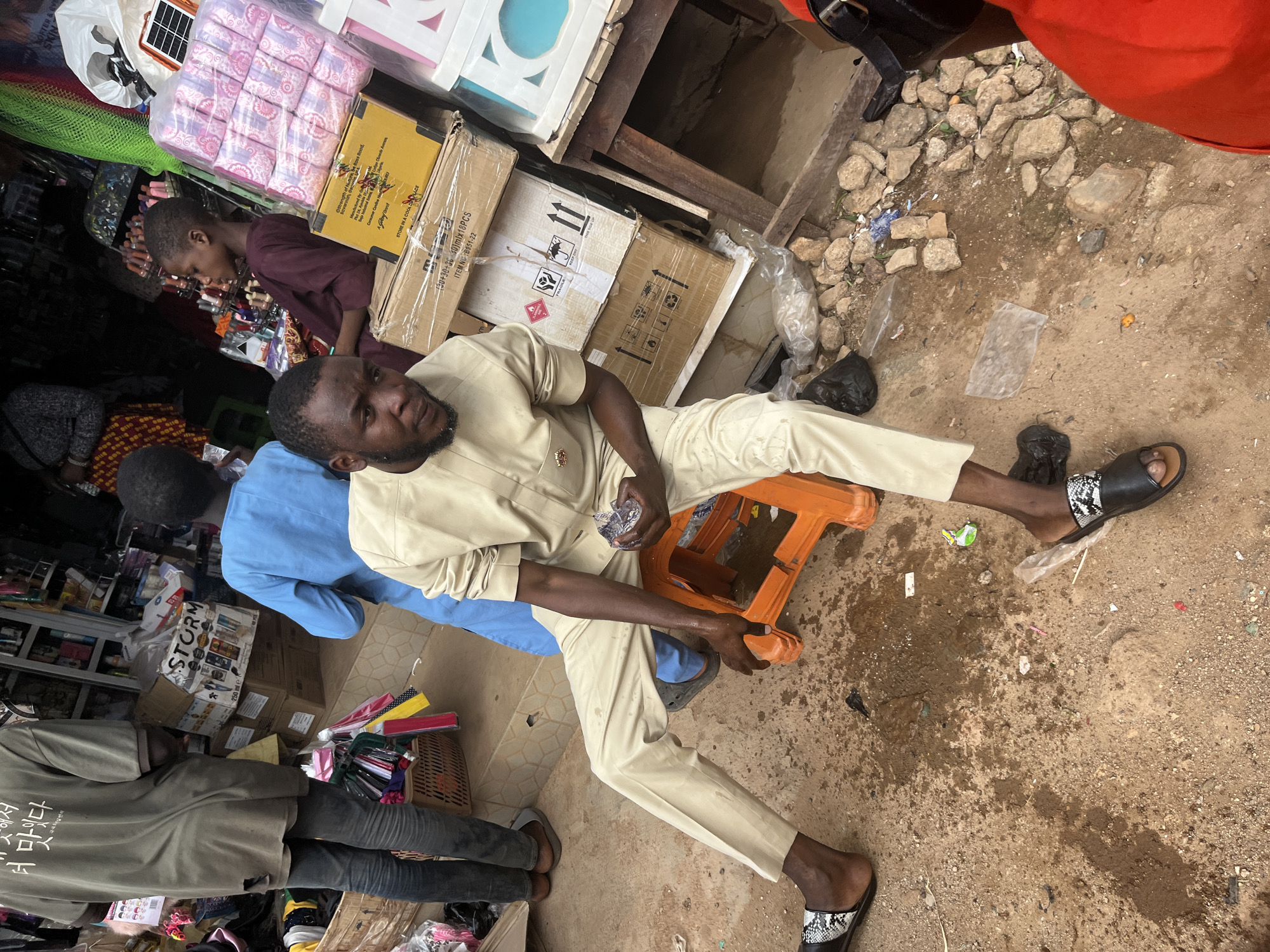 In Jos, Nigeria, a busy market scene unfolds with a focal point on a man in a light-colored traditional outfit. He is seated on an orange plastic stool, his legs crossed, and he is holding a crumpled clear plastic object in his right hand. His attire consists of a long-sleeved tunic and matching trousers, complemented by black and white patterned slide sandals. Beside him, to his right, is a partially visible person in an orange garment.
In the background, other individuals are present. To the left, a person in a faded green t-shirt and dark trousers stands near a stall filled with various items. To the right, a young boy in a dark purple garment is looking towards the right of the frame. Another person, wearing a bright blue outfit, is visible behind the man on the stool.
The environment appears to be an outdoor market, characterized by dirt and scattered stones on the ground. Several cardboard boxes are stacked in the mid-ground, some with labels and shipping markings. A basket containing colorful items, possibly fabric or accessories, is positioned to the left. Shelves in the background are stocked with products, and a white plastic bag with pink items, likely sanitary pads, is prominently displayed. The overall atmosphere suggests a bustling commercial area.