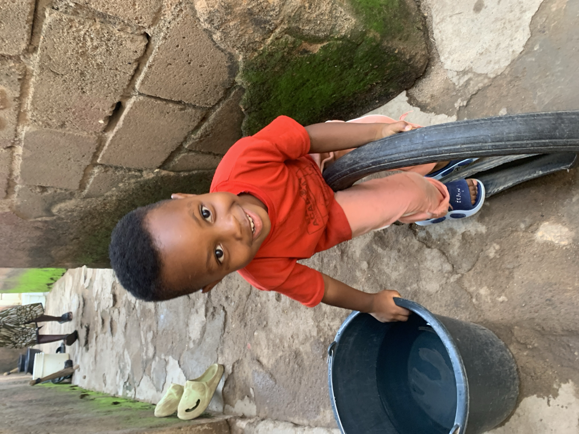 A young boy with dark skin and short black hair, wearing an orange t-shirt and light pink pants, smiles at the camera. He is holding onto a bicycle tire. To his left is a large black bucket filled with water. In the background, a person wearing a patterned skirt walks away from the camera down a narrow alleyway with rough, textured walls on either side. The ground is uneven and appears to be made of concrete. The overall scene suggests a humble, outdoor setting in a residential area.