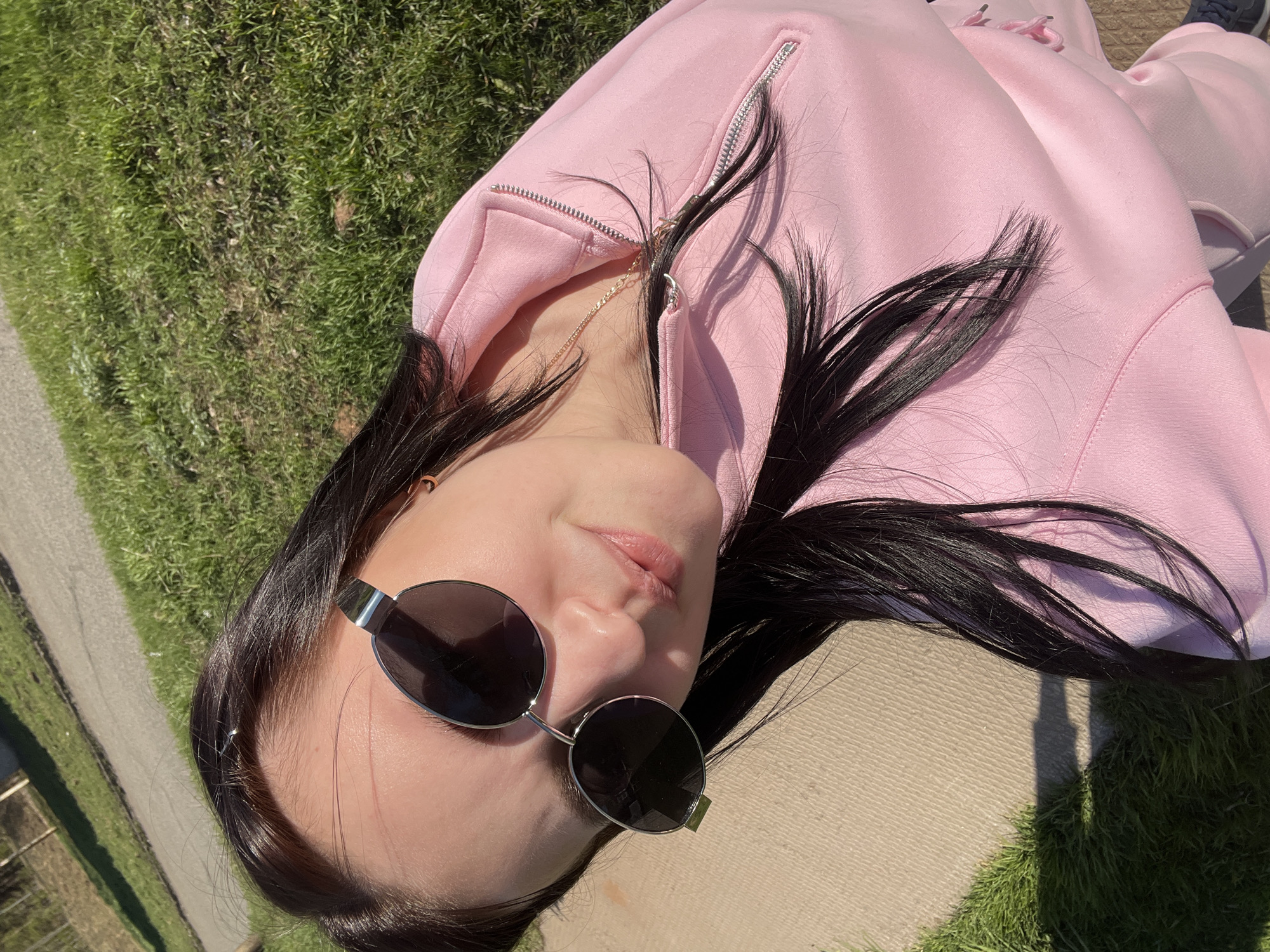 A woman with long dark hair and sunglasses is lying on a concrete surface near a grassy area in Марковский, Russia. She is wearing a pink zip-up jacket and matching pants. A gold necklace is visible around her neck. Part of the frame includes a grassy area and a glimpse of a paved path. The image is taken in daylight with strong sunlight.