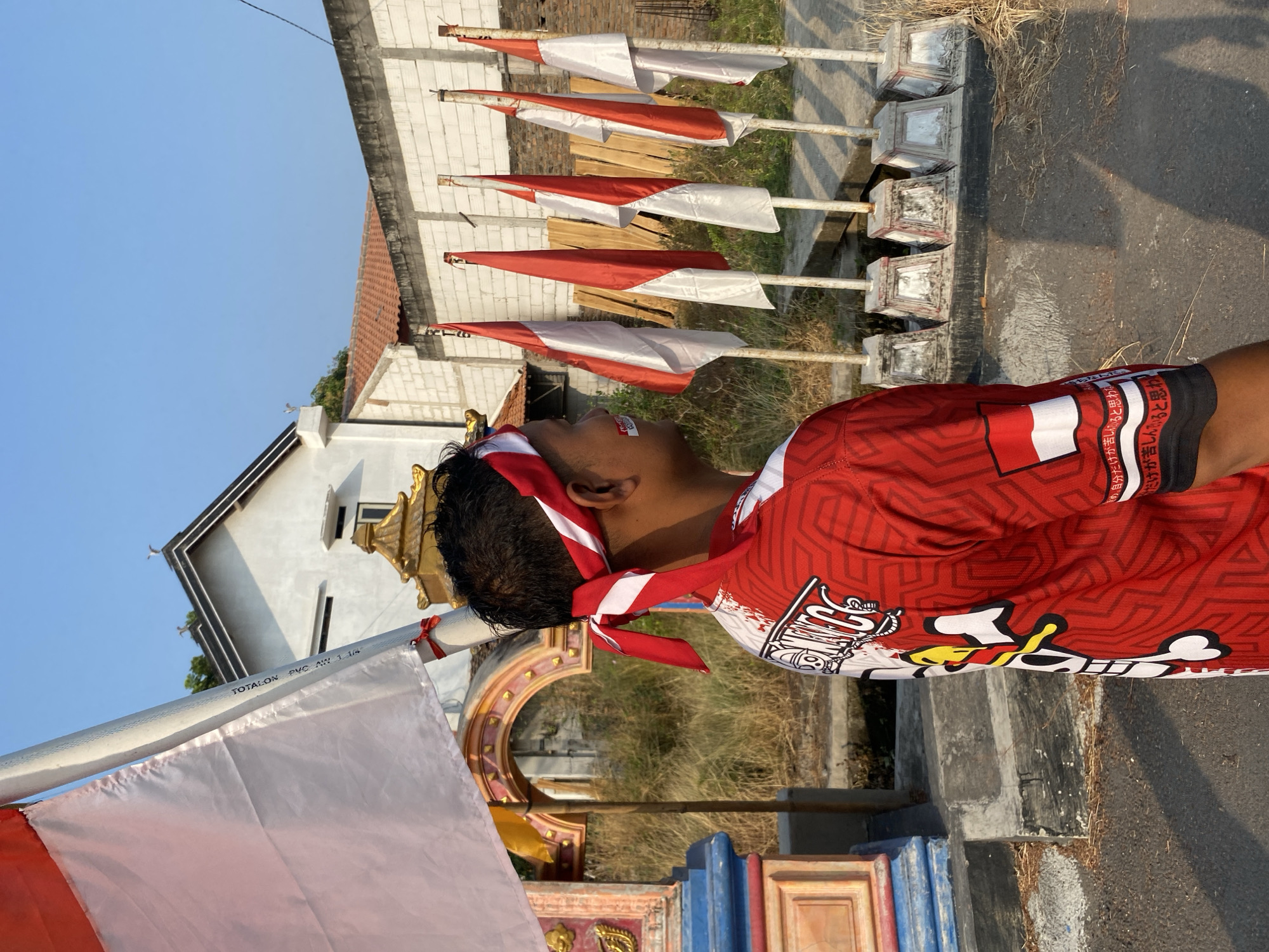 A person wearing a red and white headband and a red jersey with white accents is standing outdoors. The person has a red and white pattern on their cheek. They are looking to the right of the frame. In the background, there are several red and white flags on poles, a white building with a tiled roof, and a wall made of concrete blocks. The ground in front of the flags appears to be a paved surface or pathway. To the left of the person, there is a decorative structure with arched openings and colorful detailing, partially obscured by a large white flag on a pole. The environment suggests a public outdoor space, possibly during a celebration or event, given the patriotic imagery of the flags and headwear. The lighting indicates it is daytime, with clear skies. The location is identified as the city of Wonosari, Indonesia.