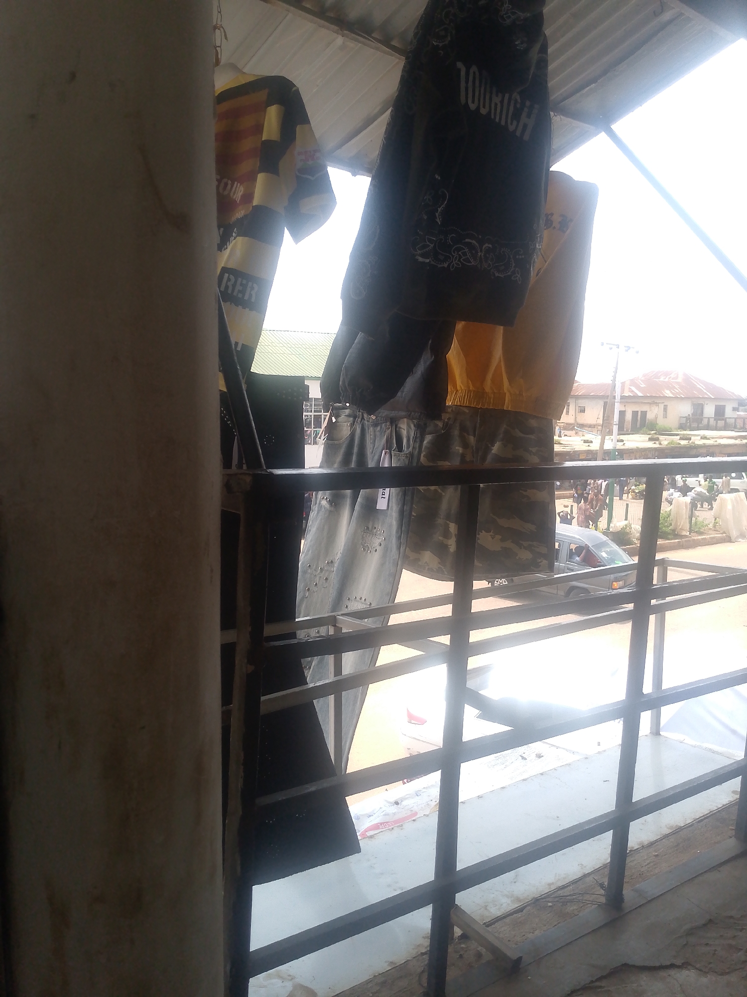 The image depicts a scene viewed through a metal railing, likely from an elevated vantage point such as a balcony or a shop's upper floor. In the foreground, a beige concrete pillar is visible on the left, with a dark metal railing running horizontally and vertically across the frame. Hanging from a chain above, several pieces of clothing are displayed.
The clothing includes a horizontally striped shirt with yellow, black, and red bands, with the word "FOUR" visible. Next to it, a dark garment with white lettering that reads "100RICH" is hanging. Below these, a pair of light-wash denim jeans with embellishments and a pair of camouflage-patterned pants are displayed.
Beyond the railing, the scene opens to an outdoor environment. A street is visible with a few people walking, and several buildings with corrugated metal roofs in the background. A grey van is parked on the street, and there are some market stalls or shops on the side. The sky is bright and appears to be daylight. The overall impression is of a market or commercial area in a town or city.