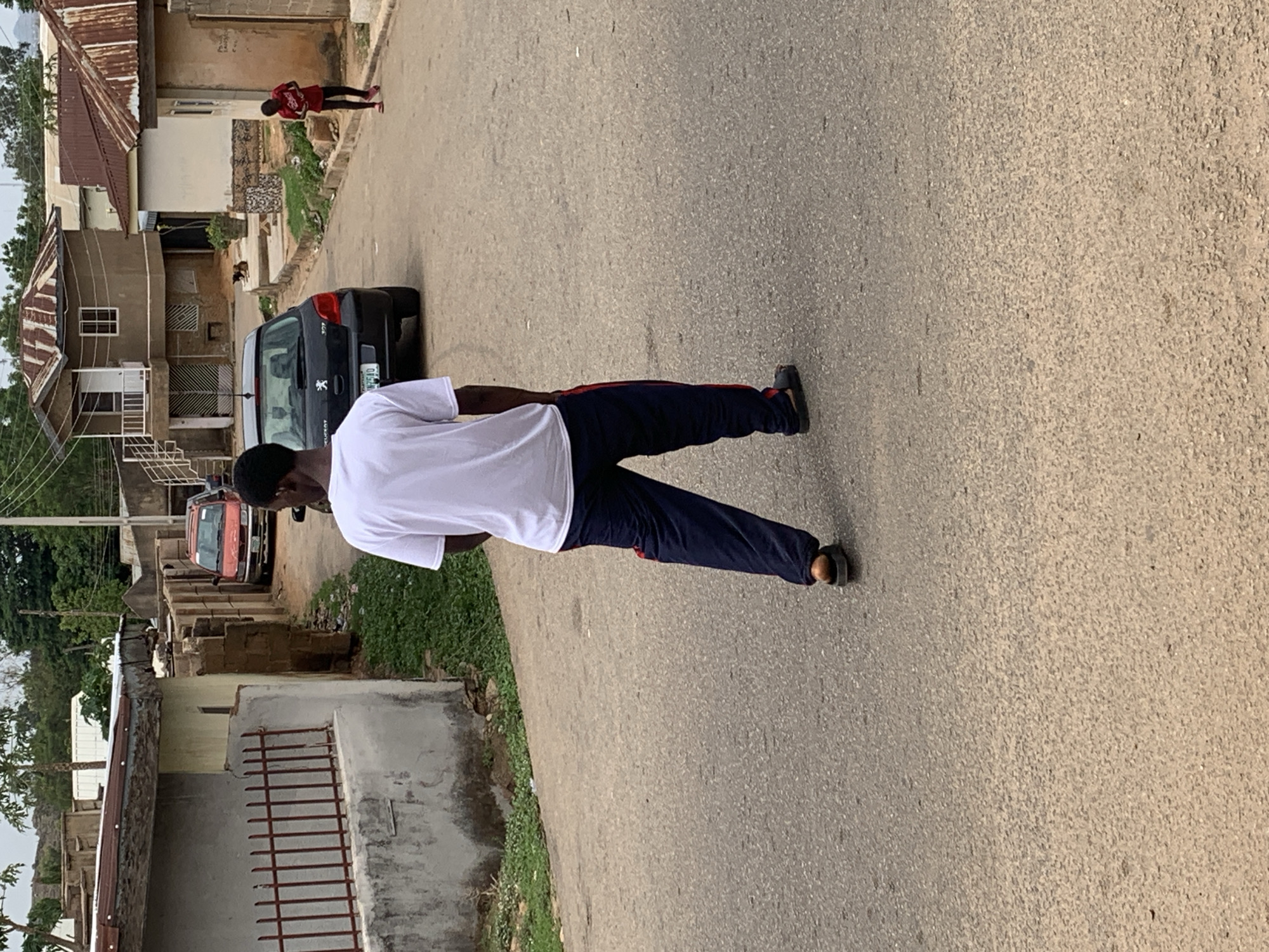 The image depicts a man walking on an asphalt road in what appears to be a residential area in Jos, Nigeria. He is wearing a white t-shirt and dark-colored pants with a red stripe. He is walking away from the camera. There is a dark-colored car parked on the side of the road. In the background, there are buildings with corrugated metal roofs, typical of residential areas in Jos. Another person is walking further up the road. The environment appears to be somewhat urban, with vegetation visible along the sides of the road. The road is lightly textured.