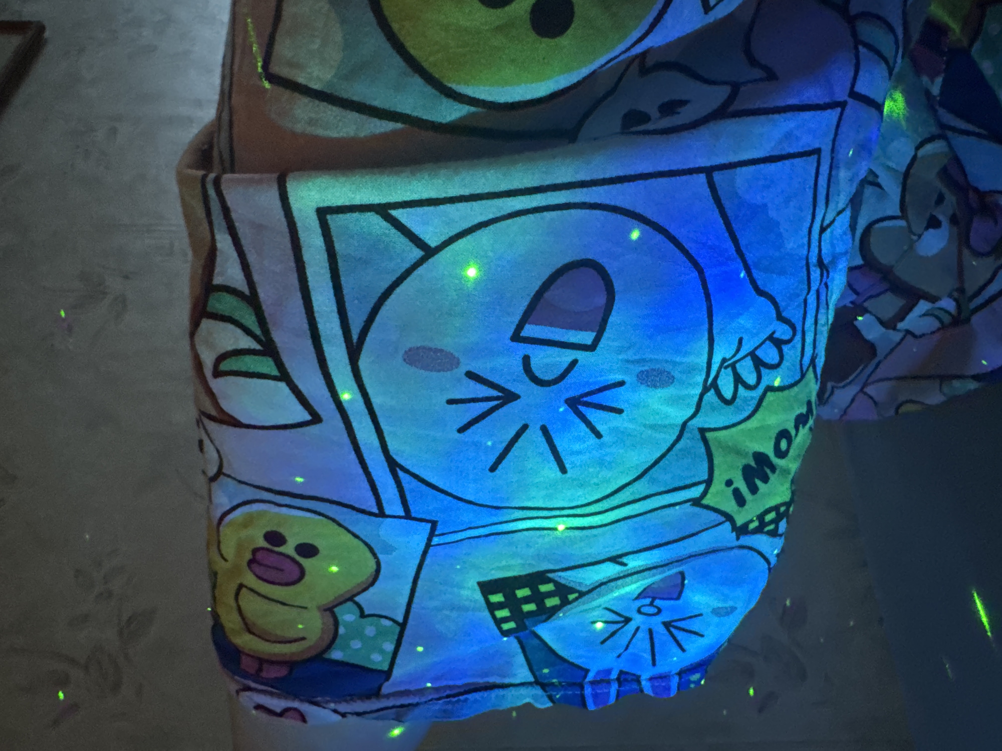 The image depicts a close-up of fabric printed with cartoon characters. Dominant colors are blue, yellow, and green. The fabric has cartoonish faces and speech bubbles. One character resembles a cat with whiskers and a wide mouth. Another character is a yellow duck with a smile. A speech bubble shows the word "imom". Tiny green specks are scattered throughout the image. The background is nondescript and out of focus. The location and city are not discernible from the image.