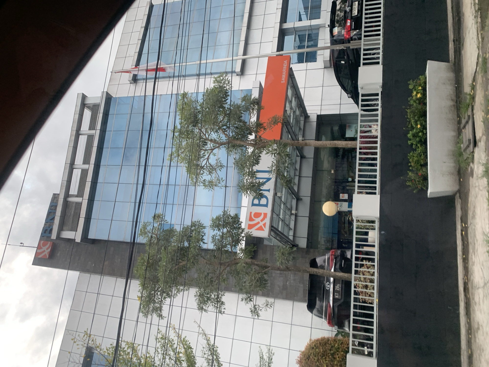 The image shows the exterior of a modern, multi-story building in Samarinda, Indonesia. The building features a glass and white panel facade. Signage indicates the presence of a Bank Negara Indonesia (BNI) branch. An orange banner displaying "SAMARINDA" is visible above the bank signage. A red and white Indonesian flag flies atop the building. Several trees are in front of the building. A black car is parked near a white fence. The sky is overcast.