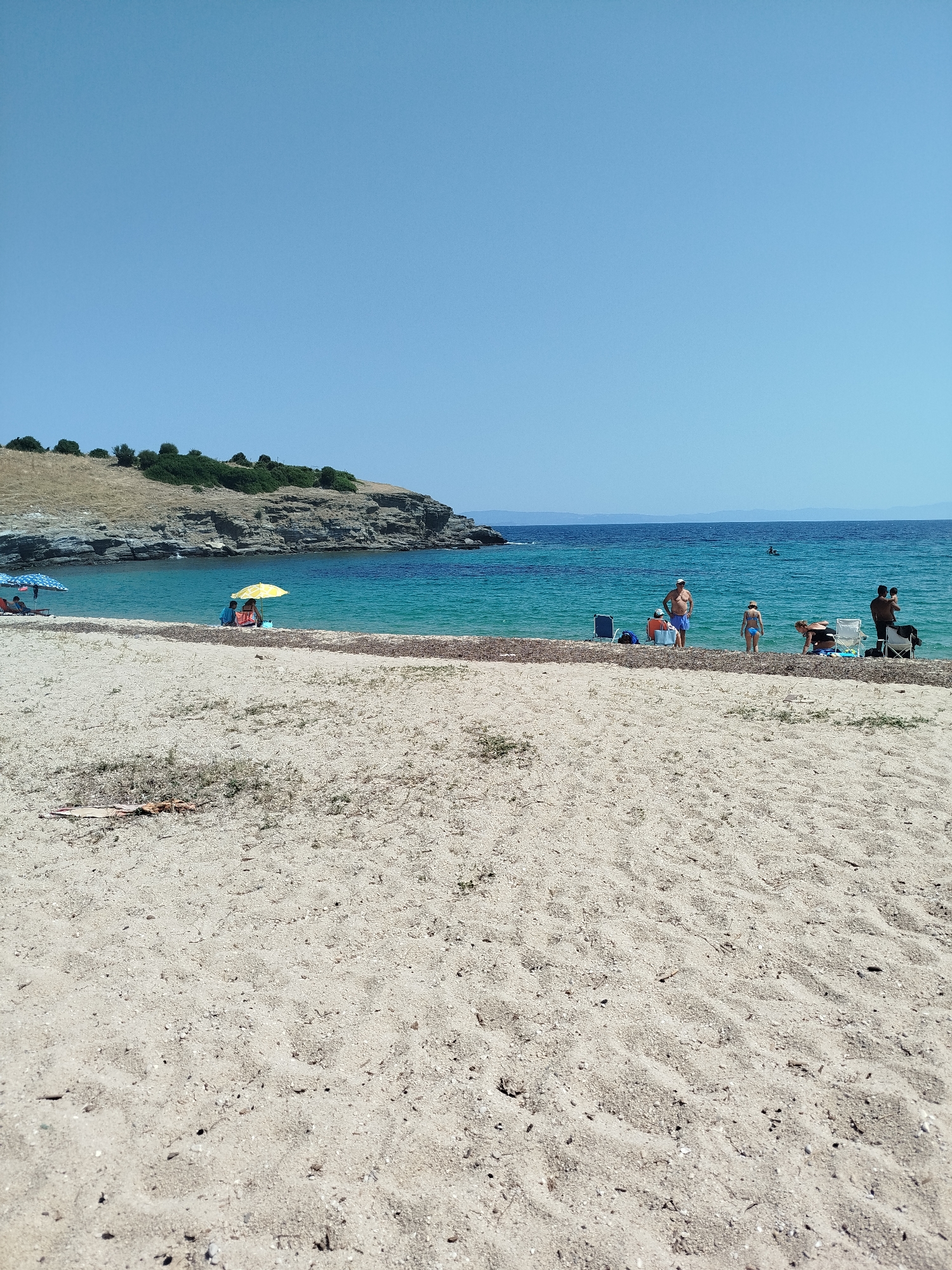 The image captures a sunny day at a beach in the Sithonia Municipal Unit, Greece. The foreground is dominated by sand, with scattered dry vegetation and debris. A line of seaweed or debris separates the sand from the shallow water. The water is a vibrant turquoise color, gradually deepening to a darker blue further out. In the middle ground, several people are enjoying the beach. On the left, two individuals are seated under a blue umbrella. Further to the right, a yellow umbrella shades two people, one of whom is looking down. Adjacent to them, a group of people stands near the water. One man in blue shorts is facing the camera, with a cooler and bags beside him. A woman in a blue bikini is standing in the water, and another woman is crouched down. On the far right, a man holds a baby. In the background, a rocky coastline with sparse vegetation rises from the sea. The sky is a clear, bright blue.