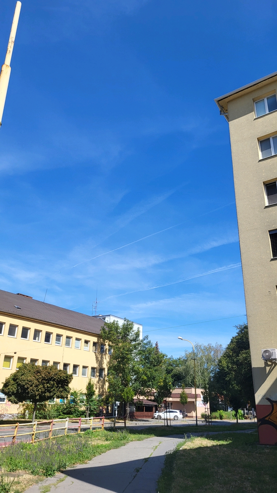 The image depicts a daytime scene on a street in Prešov, Slovakia. The sky is a vibrant blue with several white contrails streaking across it. On the right side of the frame, a light-colored apartment building occupies a significant portion of the view, with visible windows and an outdoor air conditioning unit mounted on its exterior wall. In the mid-ground, a long, yellow apartment building is visible with numerous windows. In front of this building, a colorful metal fence separates the sidewalk from a patch of grass and wildflowers. Several trees are scattered throughout the scene, some with dense foliage and others more sparse. A white car is parked near a small, covered outdoor seating area. A tall, light-colored pole, possibly a lamppost or part of a structure, is visible on the far left side of the image. The ground is paved with concrete, and some areas show signs of wear and grass encroaching.