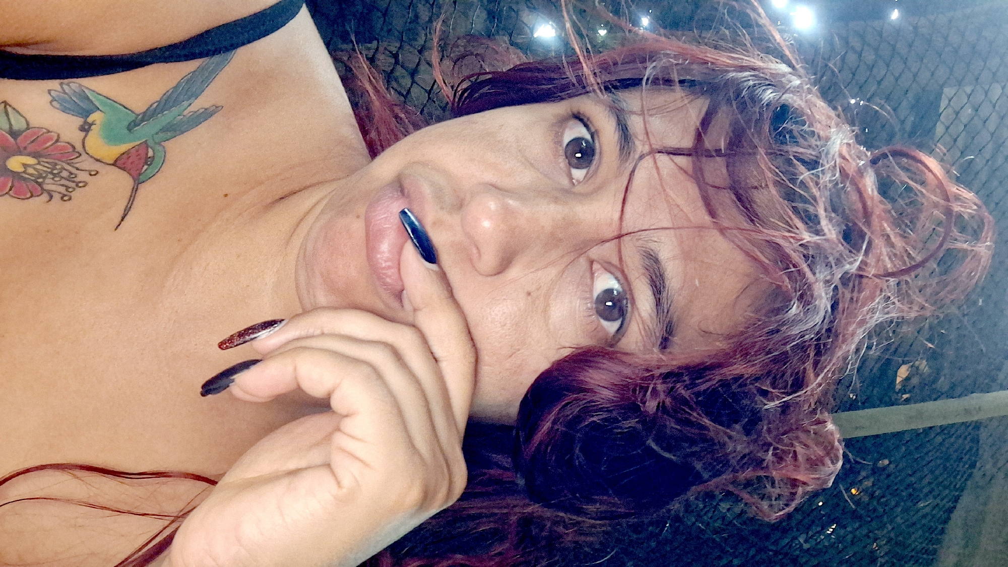 A woman with auburn hair pulled into a messy bun, and some strands framing her face, looks directly at the camera. She has brown eyes and is wearing black nail polish on her right thumb, which is near her mouth. Her left hand, adorned with dark, glittery nail polish, rests on her chin. She is wearing a black strap on her left shoulder, and a tattoo of a hummingbird in flight with outstretched wings, colored green, yellow, and red, is visible on her upper chest. Below the hummingbird, a portion of another tattoo featuring a pink and red flower is visible. The background is dark and out of focus, with some blurry lights suggesting an outdoor setting at night. The scene is located in Vallecito, Venezuela.