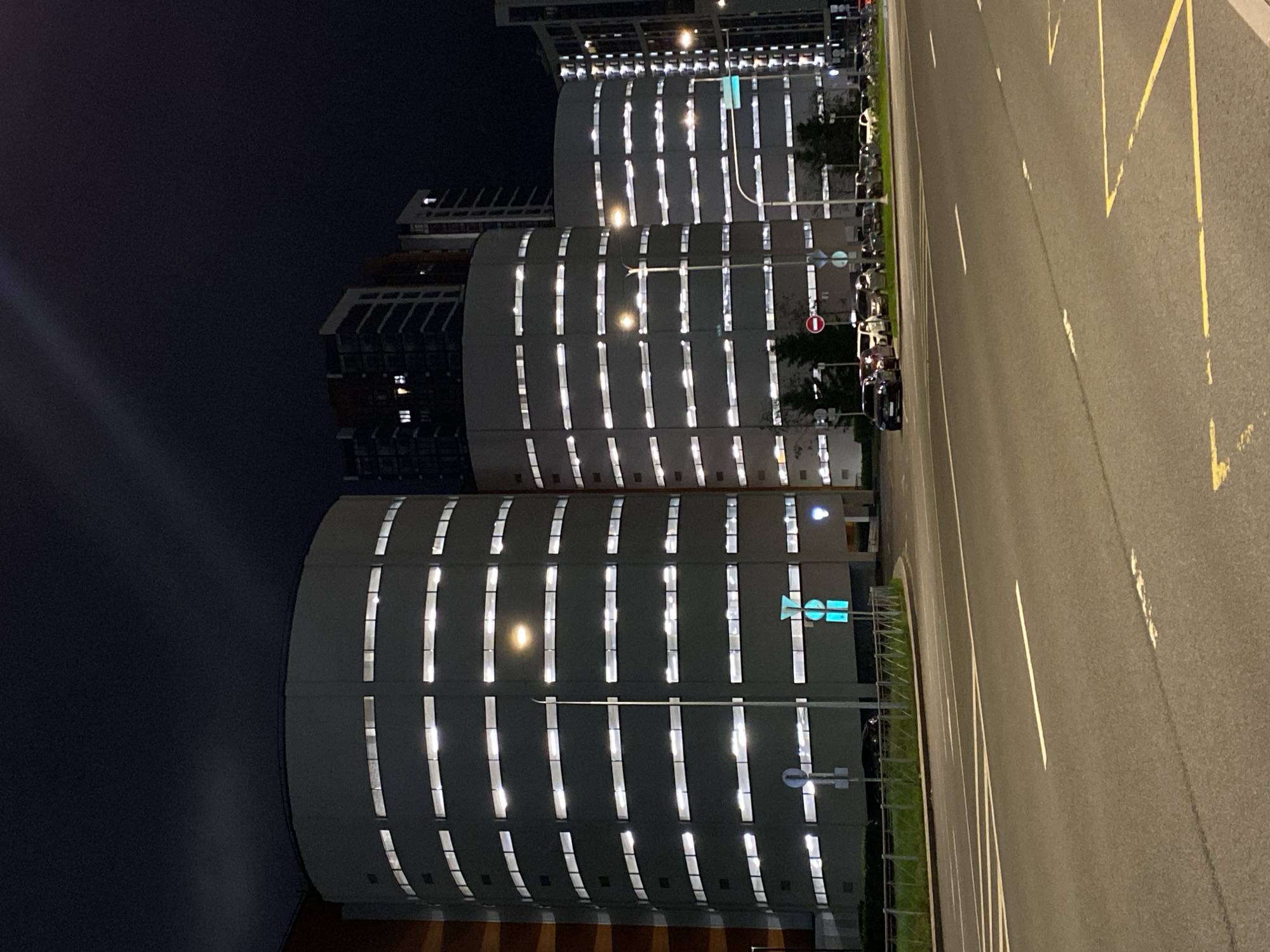 At night, the scene shows several cylindrical buildings with rows of illuminated windows next to a wide road. The road has yellow zigzag lines and white stripes. Cars are parked along the roadside. A traffic signal is visible. The location is Saint Petersburg, Russia.