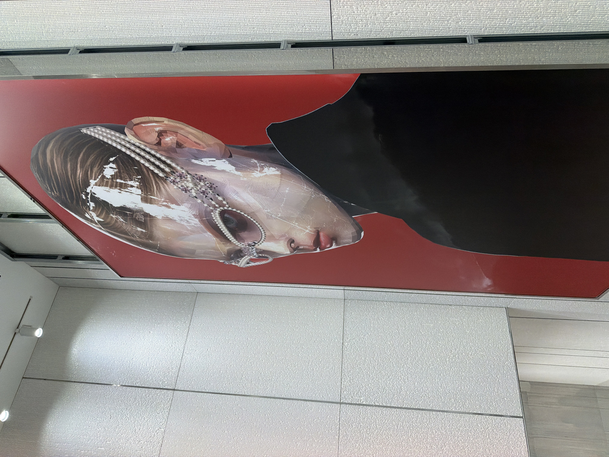 A large, vertical poster featuring an artistic depiction of a person's head and shoulders against a red background is mounted on a textured, light-colored wall. The poster displays a stylized, almost alien-like face with metallic-looking skin, wearing elaborate pearl and jewel-encrusted eyewear. The person has dark hair pulled back and is wearing a black garment. The wall has a pattern of fine vertical ridges, divided into rectangular sections. The environment appears to be indoors, possibly a public space or a gallery, with lighting fixtures visible on the ceiling. This poster is located in the Chuo City, Japan.