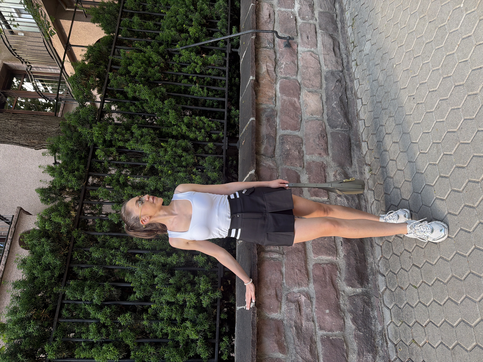 A young adult female stands outdoors on a paved sidewalk in Sofia, Bulgaria, positioned in profile and looking to the right. She wears a white square-neck tank top, a black pleated mini-skirt with two horizontal white stripes on the wearer's left side, and white and silver chunky athletic sneakers. Her dark hair is pulled back into a high ponytail secured with a gold-colored accessory, and she wears light-tinted oval sunglasses with thin gold frames. Her left forearm and hand rest on the dark capstone of a low stone wall constructed from reddish-brown and grey rough-hewn stones. Her right hand holds an olive green shoulder bag with a gold-colored clasp. Behind the wall, a black metal fence is partially visible through dense, dark green evergreen shrubs. In the background, portions of a light-colored building with brown-framed windows and a metal-railed balcony are discernible, along with a thick tree trunk on the far left. The foreground consists of grey hexagonal interlocking pavers. The scene is illuminated by strong natural daylight, indicating daytime.