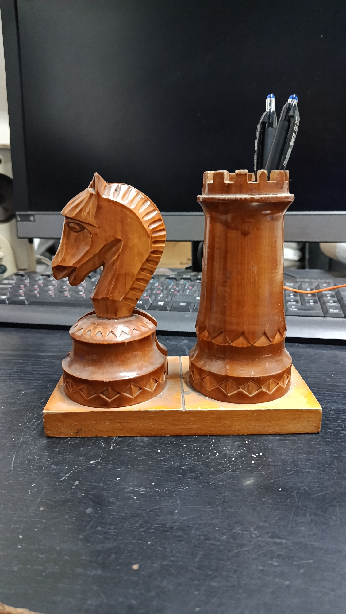 The image depicts a wooden desk organizer shaped like chess pieces. On the left is a knight, carved with intricate detail on its mane and face. To its right is a rook, which functions as a pen holder, with two black pens with blue tops inserted. Both chess pieces are mounted on a rectangular wooden base. The organizer is placed on a dark, textured surface, likely a desk. In the background, a computer monitor and a keyboard are partially visible, suggesting an office or home office environment. The lighting is even, and the focus is on the desk organizer. There are no people visible in the image. The scene is static, with no discernible action occurring. There is no indication of the specific location within the city of Kostanay, Kazakhstan.