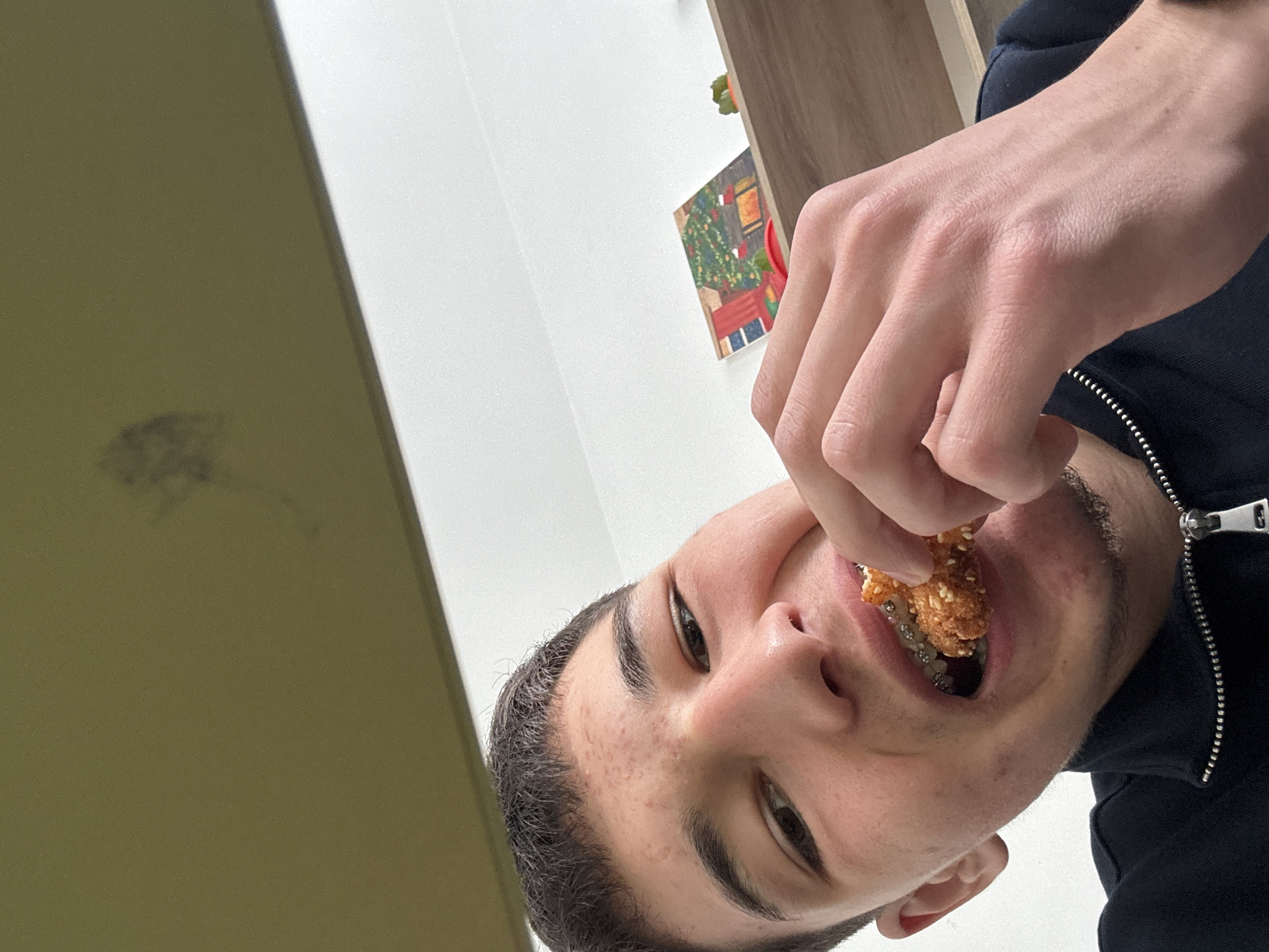 A man is holding a piece of fried food and taking a bite. He is wearing a black jacket with a silver zipper. He appears to be indoors. A calendar with a picture of a Christmas tree is on the wall. There is not enough information to determine if the image was taken in Zlatograd, Bulgaria.