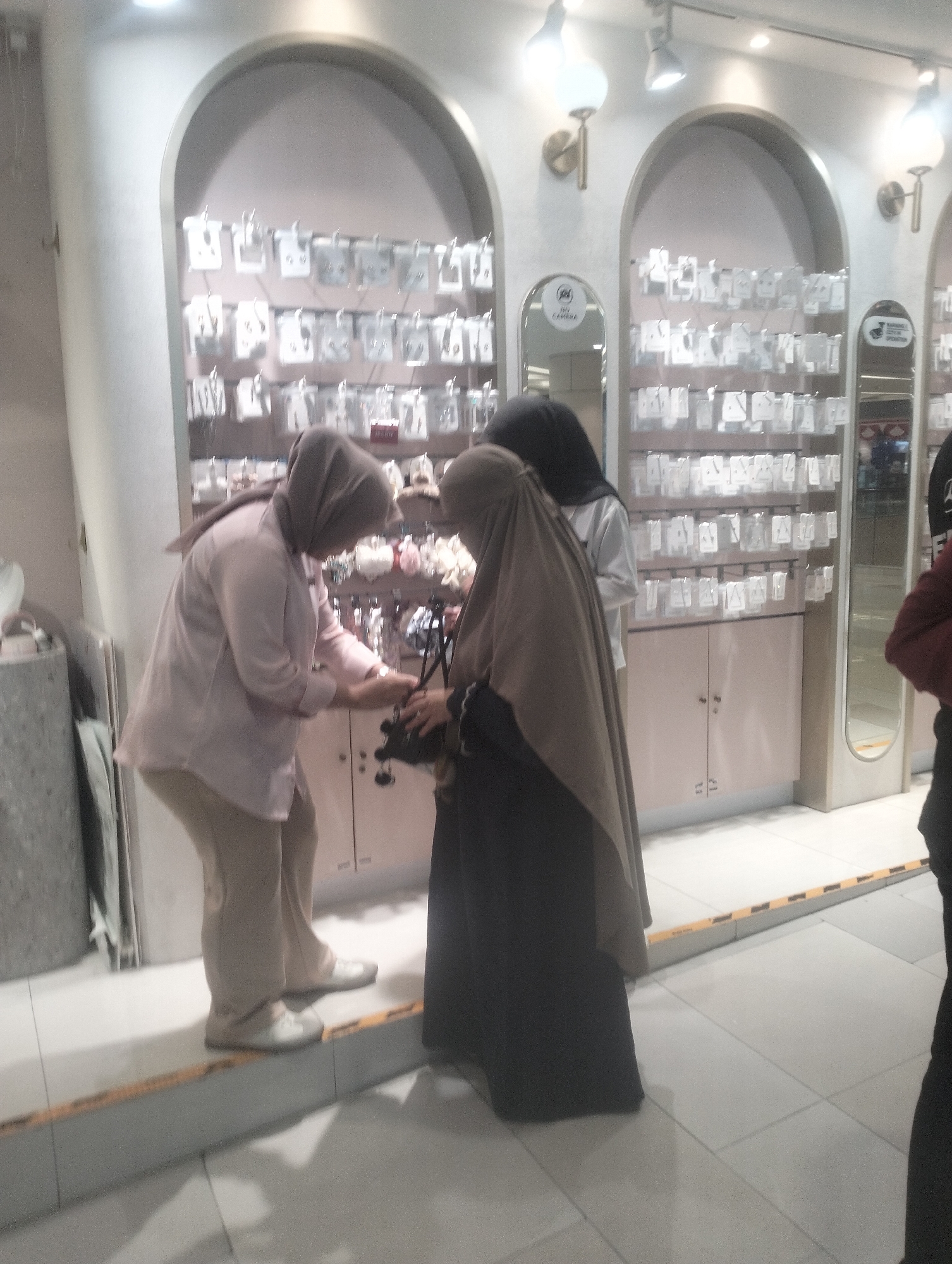 In Surabaya, Indonesia, two women are seen in a retail store, possibly a boutique or a department store, interacting near a display of merchandise. The woman on the left, wearing a light pink top and beige pants, is bending over and appears to be examining or holding a black item, possibly a camera or a bag. The woman on the right, dressed in a dark hijab and a long dark garment, is standing and observing the first woman's actions. Behind them, shelves are stocked with small, packaged items, likely accessories or jewelry, displayed in clear plastic packaging. The store features arched architectural elements and modern lighting fixtures. The floor is tiled, and there are some yellow markers on the ground, indicating a walkway or a cautionary area. The overall ambiance suggests a commercial environment focused on retail sales.