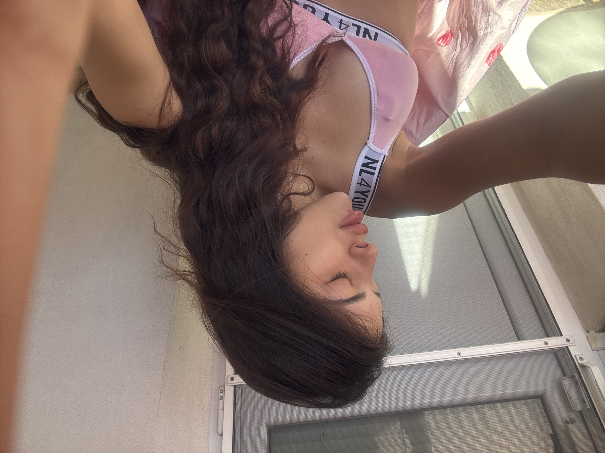 A young woman with long, dark, wavy hair is seen from the chest up in a selfie. She is wearing a light pink bra with a white band that has black lettering. Her eyes are closed, and her head is tilted slightly. Her left arm is extended towards the camera, with her elbow bent. Behind her is a window with a screen, and the background appears to be a light-colored wall. The lighting suggests it is daytime. This scene is not identifiable as being in Plovdiv, Bulgaria.