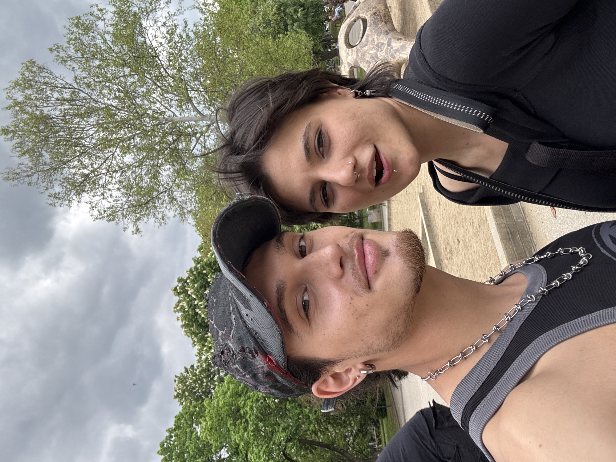 Two people pose for a selfie outdoors. The person in the foreground wears a tank top, a chain necklace, and a hat. The person behind is wearing a dark top with straps. Trees and the sky are in the background. It is impossible to identify the exact location in Sofia from the image.