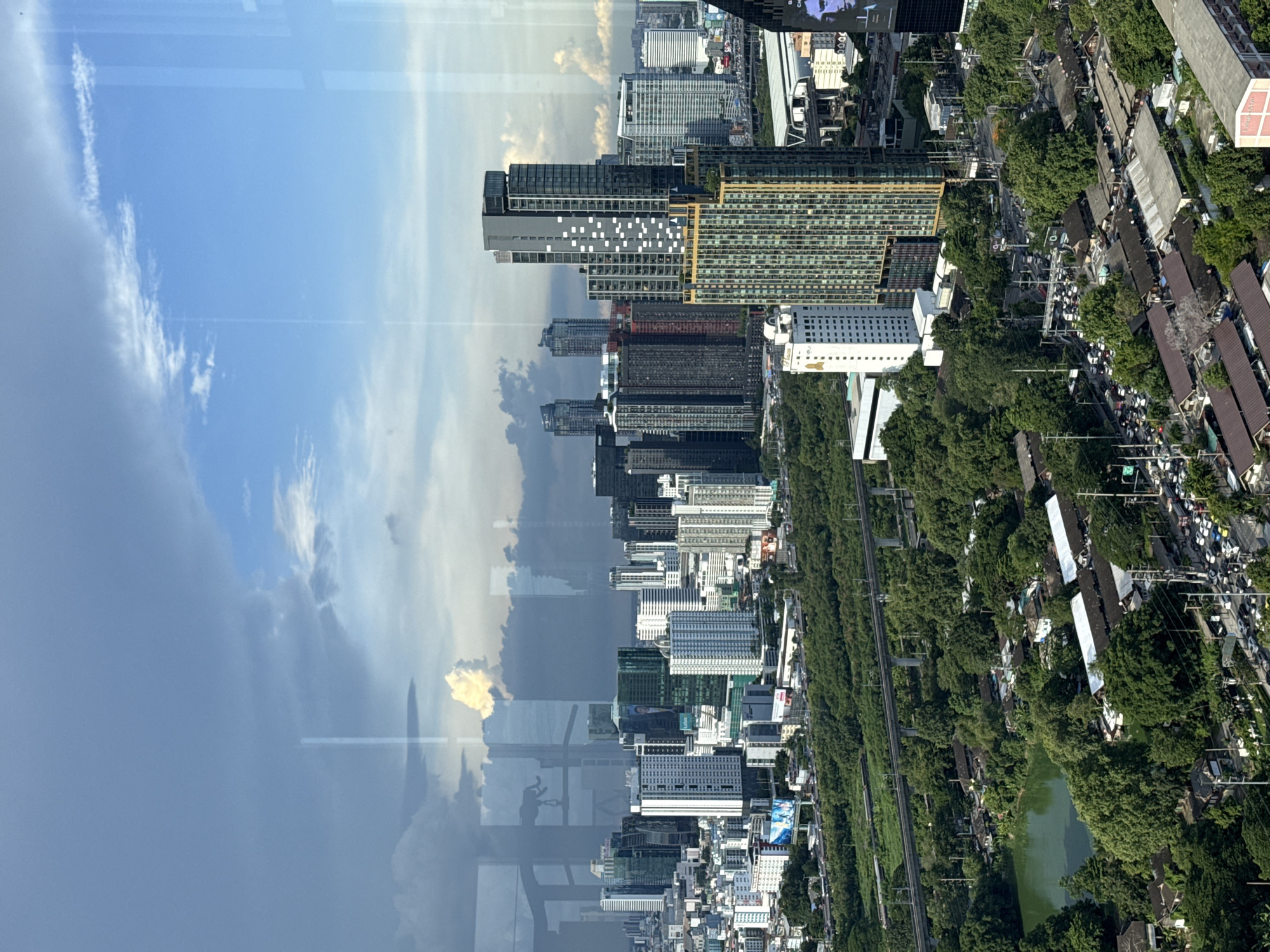 This is a vertical, high-angle shot of a cityscape, likely taken from a tall building, reflecting the sky and clouds. The scene is divided between the sky and the urban environment, with the reflection creating a mirrored effect.
On the right side of the image, we see a dense urban landscape with numerous buildings of varying heights and architectural styles, interspersed with lush green trees. Some of the buildings appear to be residential complexes, while others are taller, modern skyscrapers with glass facades. A road runs through the middle of the scene, lined with traffic, suggesting a busy urban environment. There's also a body of green water, possibly a pond or canal, surrounded by trees. The buildings and trees are densely packed, indicating a high population density. The time of day appears to be daytime, with clear blue skies and some scattered white clouds visible in the reflection.
The left side of the image is dominated by a reflection of the sky and clouds, which are dramatic and dynamic, with some areas of bright sunlight and darker, more ominous clouds. The reflection suggests the photo was taken through a window, and the angle of the shot and the reflections indicate that the image is rotated 90 degrees counter-clockwise from its natural orientation.
The overall impression is one of a bustling, sprawling metropolis, with a clear contrast between the man-made structures and the natural elements of the trees and sky. The sheer density of buildings and traffic points to a major city, and the presence of distinct architectural styles could be indicative of different development phases within the city.