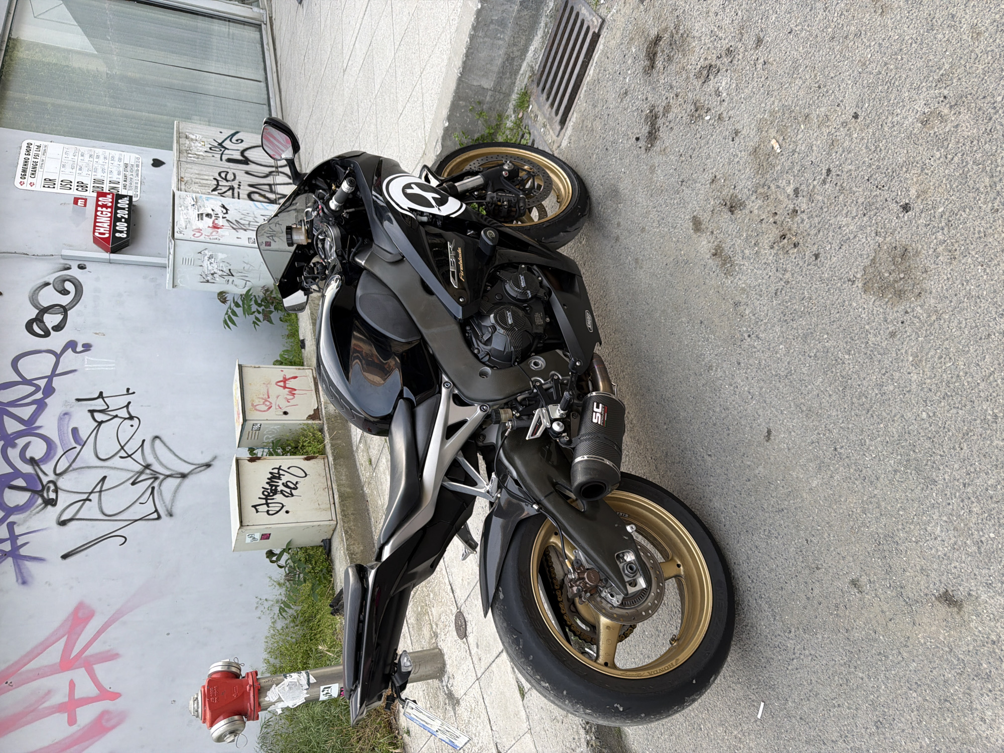 The image captures a black Honda CBR Fireblade motorcycle parked on a sidewalk. The motorcycle features gold-colored rims and an SC Project exhaust pipe. Graffiti is visible on the adjacent wall and electrical boxes. A red fire hydrant stands near the motorcycle. A sign for a currency exchange bureau, "CHANGE FSI Ltd.", is posted nearby, displaying exchange rates for EUR, USD, and RUB, indicating that the location is likely in Bulgaria. Additionally, the sign advertises "CHANGE 30" with hours of operation from 8:00 to 20:00. A metallic grid drain is nearby. The location is highly likely to be in Burgas, Bulgaria based on the text on the sign.