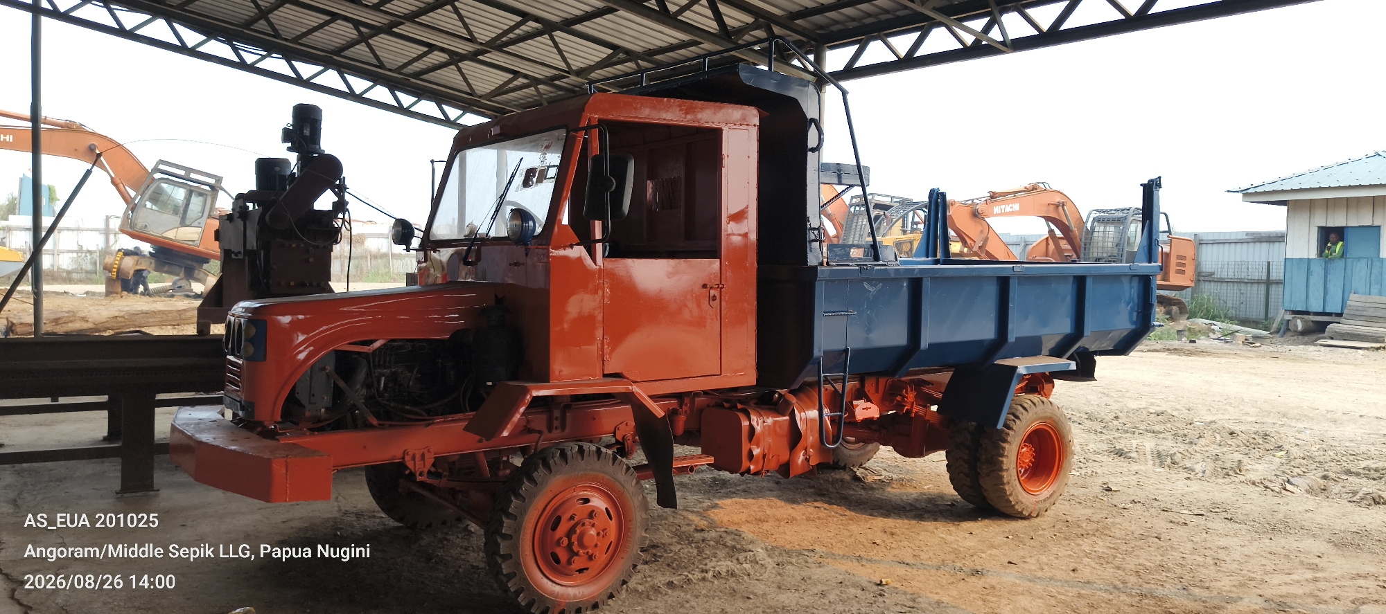 The image shows an orange and blue dump truck parked under a metal roof. The truck has a red cabin with large windows and a blue cargo bed. The truck's tires are large and rugged, and the wheels are painted orange. In the background, there are several orange excavators, some construction materials, and a small blue and white building. A person wearing a yellow vest is visible in the window of the building. The ground is dirt and appears to be a construction or industrial site. The text overlay at the bottom left of the image indicates the location as "Angoram/Middle Sepik LLG, Papua Nugini" and the date as "2026/08/26 14:00".