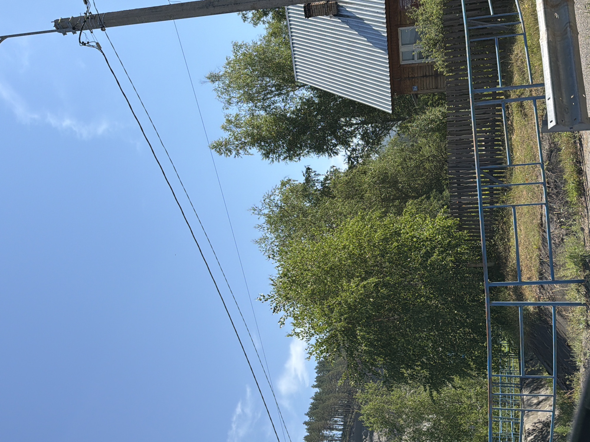 The image depicts a clear blue sky with scattered clouds over a rural landscape. A concrete utility pole with power lines stretches diagonally across the upper right portion of the frame. Below the power lines, a dense collection of deciduous trees with vibrant green foliage dominates the mid-ground. To the right of the trees, a wooden house with a corrugated metal roof and a visible chimney is partially obscured. A rustic wooden fence runs horizontally across the middle of the image, separating the foreground from the background. In the foreground, a blue metal railing runs along what appears to be a pathway or embankment, with a grey metal barrier in the lower right corner. The scene is set in the city of Uznezya, Russia, suggesting a tranquil, natural environment.