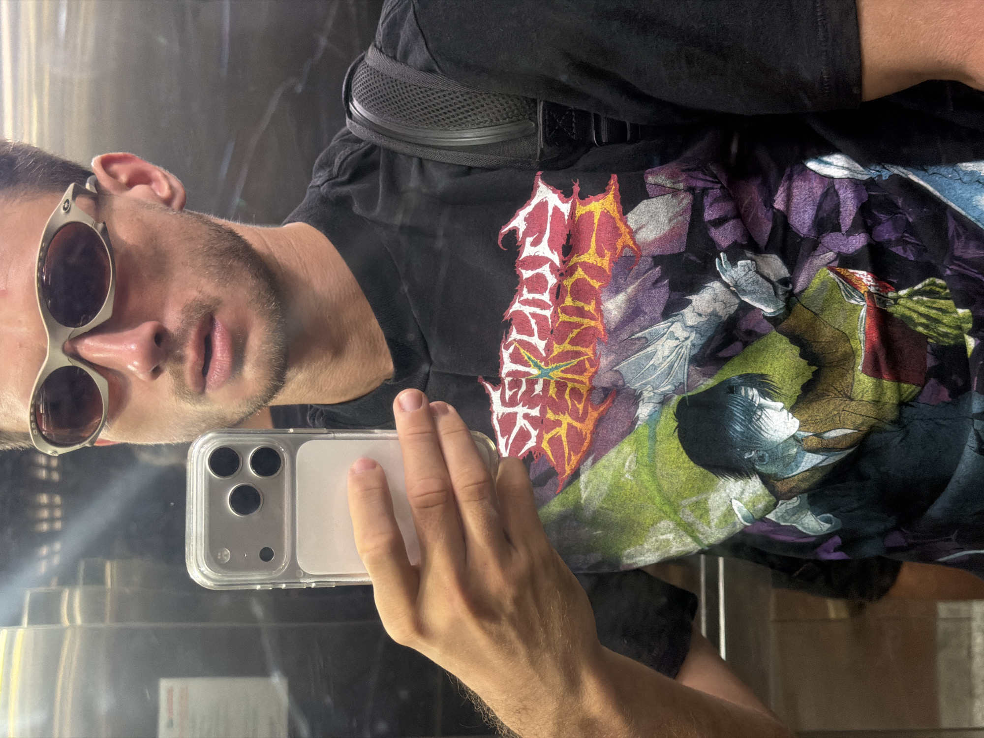 A man is taking a selfie in what appears to be an elevator. He is wearing sunglasses with silver frames, a black t-shirt with a graphic print, and a black belt with a mesh design. He is holding a silver smartphone with a clear case in his left hand. The background shows reflective elevator walls. There is no indication of Đà Nẵng, Vietnam in the image.