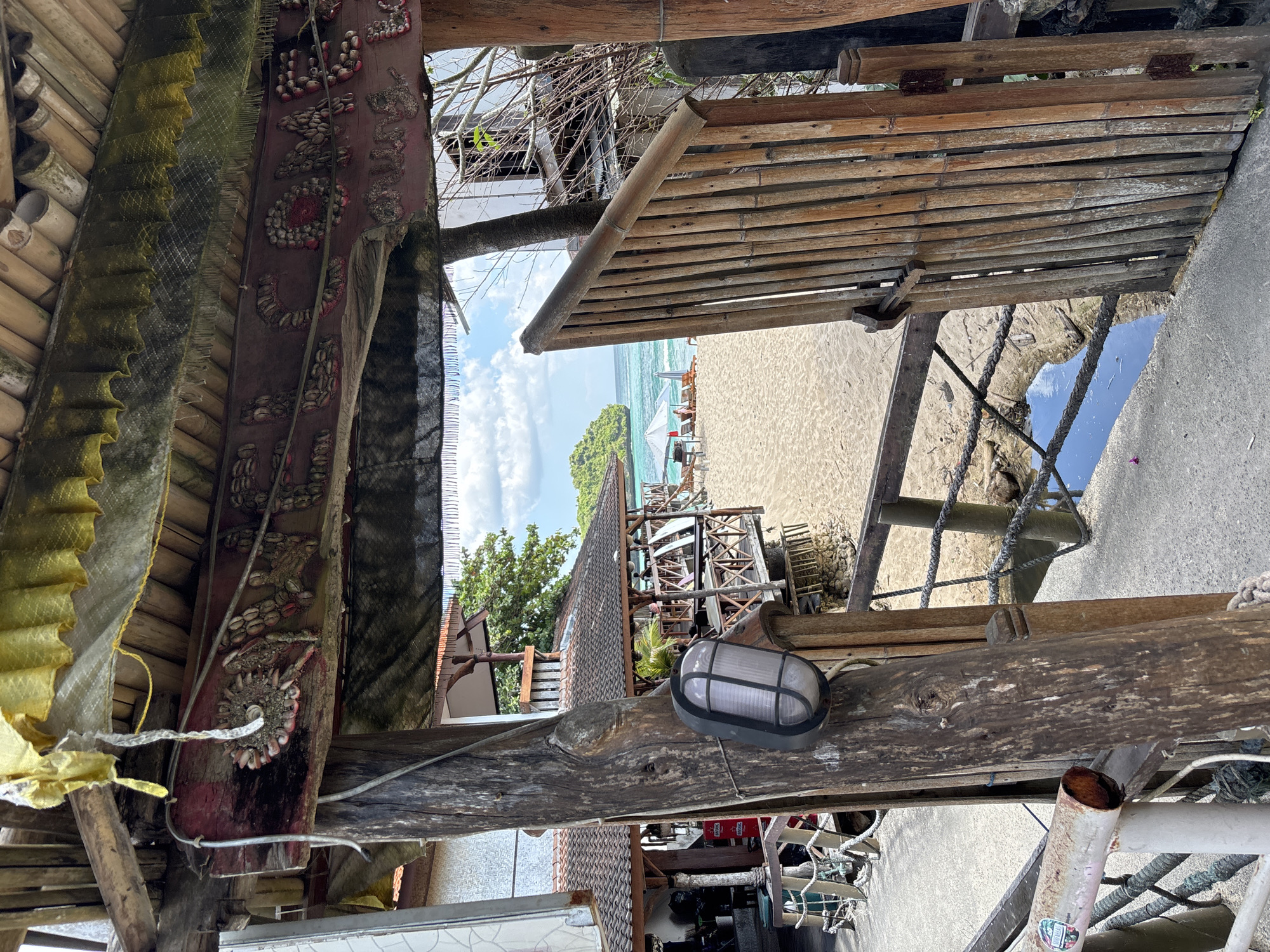 The image displays a detailed view through a rustic, open gateway constructed from rough-hewn timber and bamboo, looking out onto a sandy beach and the ocean. The horizontal wooden beam spanning the top of the gateway is adorned with shells arranged to spell "WELCOME" and a second, partially visible line of text, "Nunggal." Above this beam, a ruffled yellow fabric valance and dark mesh hang beneath a ceiling of bamboo poles.
On the left side of the gateway, a robust wooden post supports a grey, oval-shaped outdoor light fixture. Behind this post, a multi-tiered building with a red tiled roof and dark wooden railings is visible, suggesting a restaurant or hospitality establishment. Red signs bearing the "BINTANG" logo are discernible on the building's exterior.
On the right, a bamboo slatted gate, featuring a simple wooden latch, is partially open, revealing more of the sandy environment beyond.
The immediate foreground consists of a concrete walkway with rope railings on the left. A narrow channel of shallow water, possibly a tidal pool or small stream, runs between the concrete path and the main expanse of the sandy beach.
On the sandy beach, several white sun umbrellas are erected, accompanied by lounge chairs. A few individuals are observed relaxing under one of these umbrellas in the distance.
Beyond the beach, the turquoise ocean extends under a sky with scattered white clouds. A prominent, green, rocky island or headland is visible in the left mid-distance. The overall scene depicts a tropical beach setting within a resort or coastal establishment in Kuta Selatan, Indonesia, potentially Nunggalan Beach, given the visible text.