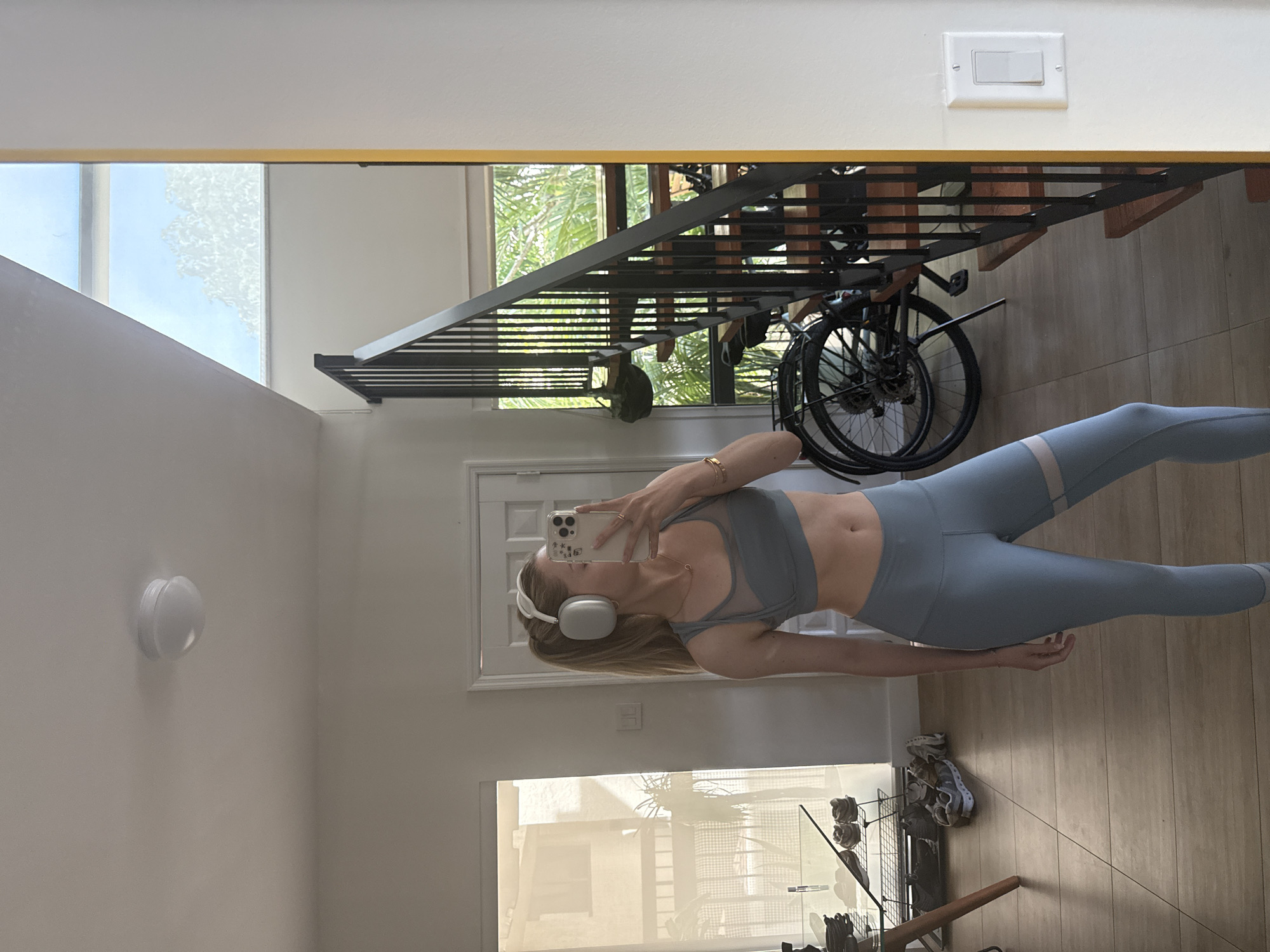 A blonde woman wearing a light blue sports bra and matching leggings, white headphones, and a necklace stands in front of a mirror, taking a selfie with her smartphone. She is in an indoor setting with wooden floors and a modern staircase visible in the reflection. The room appears to be a home or apartment. The image is likely not from Miami, United States of America, as there are no identifiable landmarks or characteristics suggesting that location.
