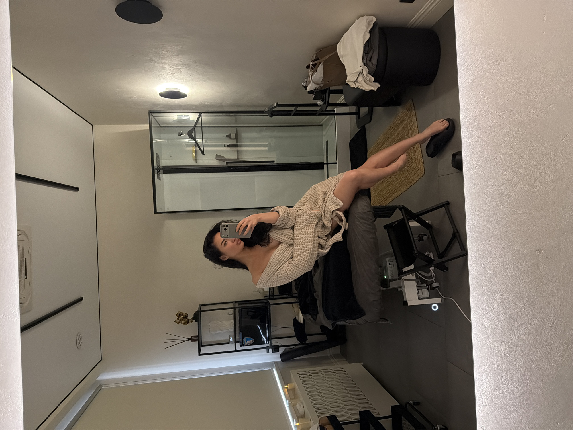 A mirror selfie shows a woman in a bathroom. She is lying on a gray chaise lounge, wearing a light-colored knit robe and holding a phone in her left hand, presumably taking the picture. Her dark hair is loose. A black folding chair is next to the chaise lounge, along with electronic devices and wiring. Behind the chaise lounge is a black metal shelving unit. The bathroom has a shower stall with a glass enclosure, a black ottoman with towels and clothes, and a small woven rug. There is a radiator near the bottom of the frame. The walls are light-colored, and there are ceiling-mounted light fixtures. It is not possible to determine the location of the city of Moscow, Russia, from the given image.