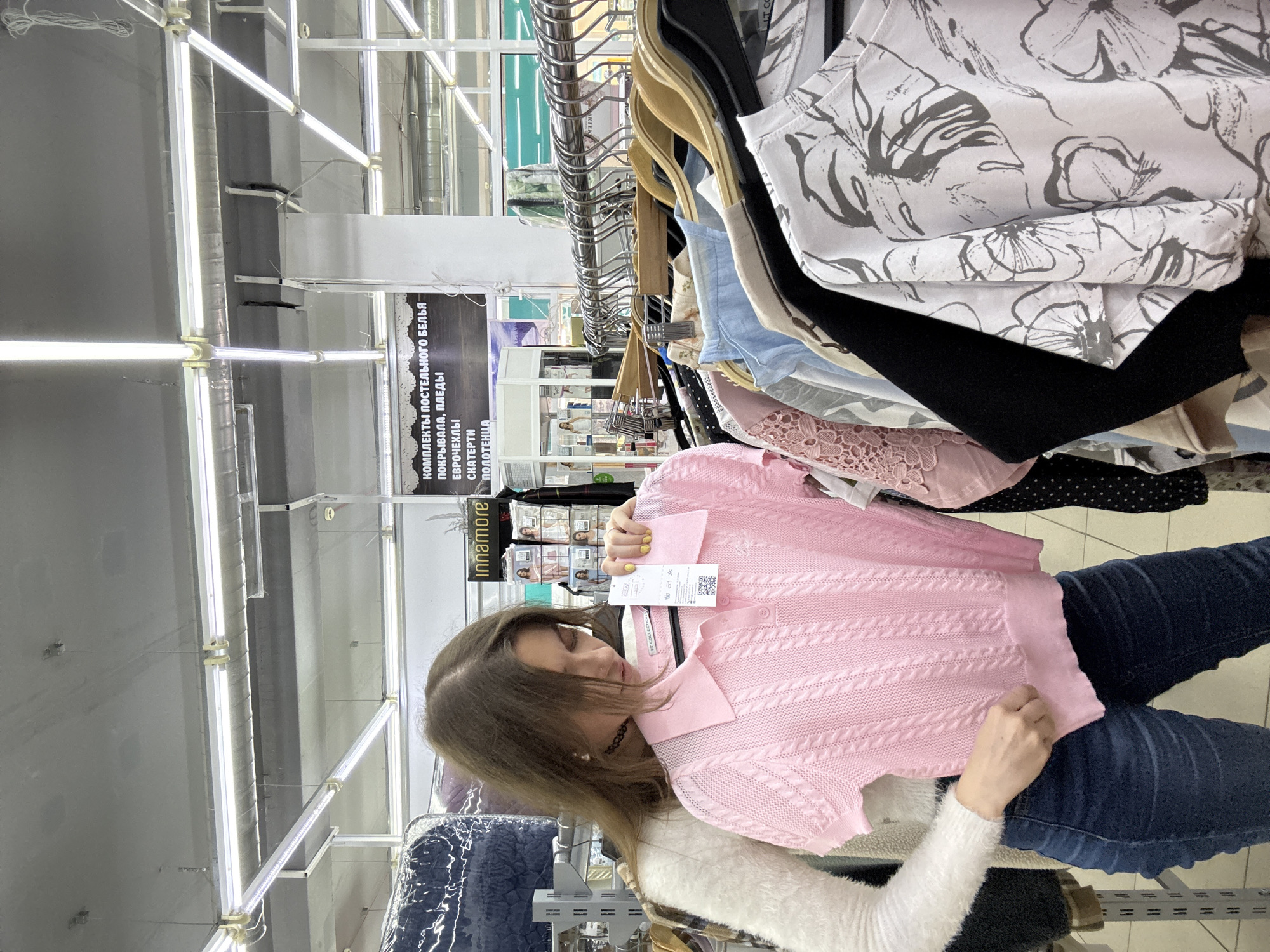 A woman with long brown hair is holding a pink polo shirt with a cable knit pattern in a retail store. She is wearing blue jeans and a white sweater with a fuzzy texture. The shirt has a price tag attached to it. In the background, there is a rack of clothing with various items, including blouses with floral patterns and black tops. A sign with Russian text is visible, indicating the store sells bedding, blankets, tablecloths, and towels. The sign also has the brand name "Innamore" on it. The location is a retail environment, likely in the city of Chaikovsky, Russia, based on the Cyrillic text. The lighting is bright and the scene appears to be indoors.