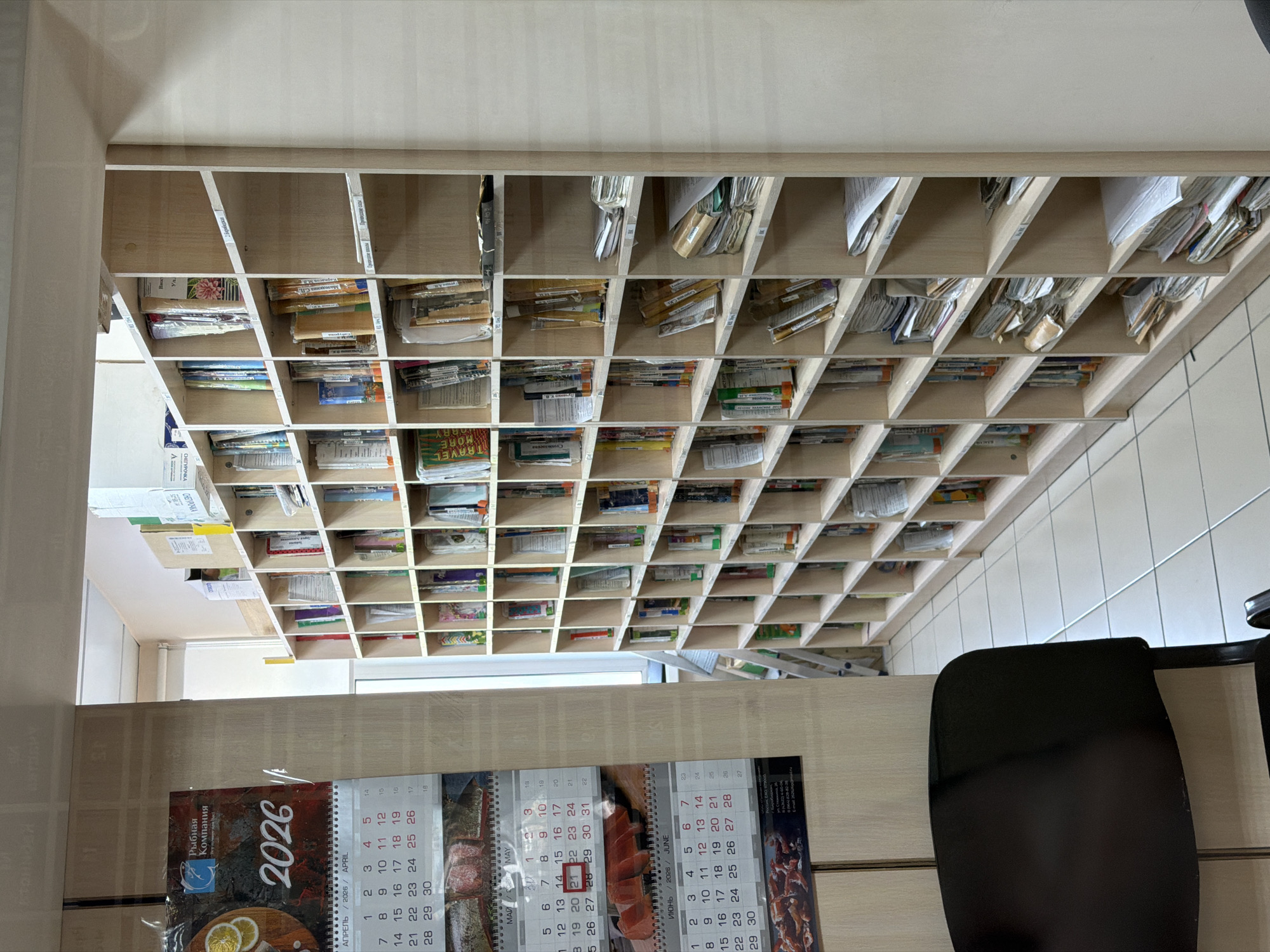 The image shows an office setting, with a large wooden shelving unit filled with various papers, documents, and small packages. The shelving unit appears to be the primary organizational system in the room. Below the shelving unit is a desk, on which a calendar for the year 2026 is visible. The calendar has the name of a company on it that translates to "Fishing Company." A black office chair is placed near the desk. The environment looks like a small office space, likely in Birobidzhan, Russia, given the name of the company on the calendar and the language of the text.