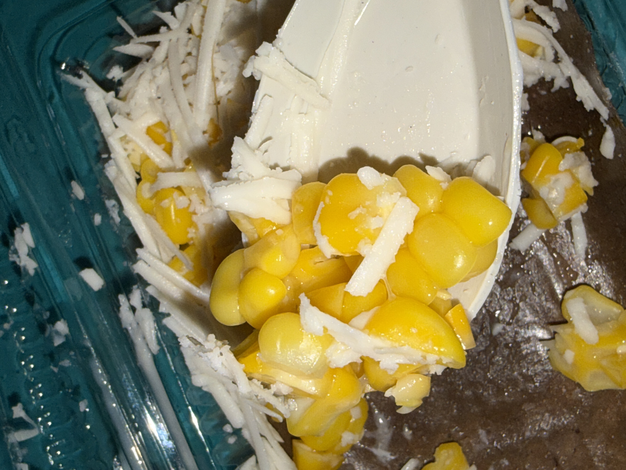 A close-up, overhead shot shows a white plastic spoon filled with corn kernels and shredded coconut. The spoon rests on a brown surface, possibly a table or counter. The corn is bright yellow and appears fresh. The shredded coconut is white and fluffy, mixed in with the corn. In the background, a teal-colored plastic container with a ribbed texture is visible, also containing corn and coconut. The lighting is somewhat dim, with some reflections on the plastic surfaces. The image does not depict any identifiable landmarks or scenes from Tupah, Indonesia.