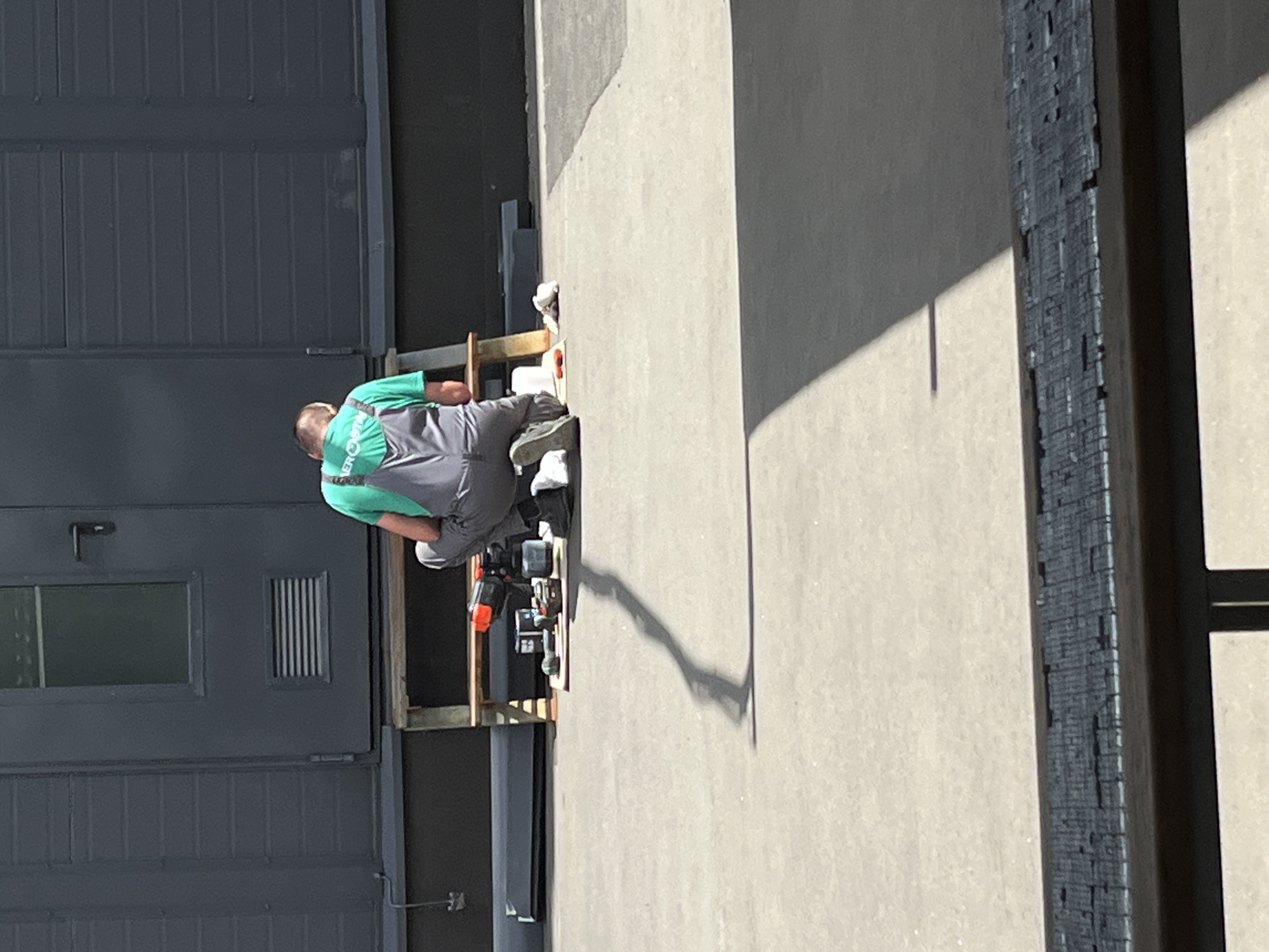 A male individual wearing a green t-shirt and grey overalls is kneeling on a concrete surface. He appears to be working with tools laid out on a wooden plank placed in front of him. The tools include what looks like a cordless drill, batteries, and other equipment. Behind the individual is a dark grey, industrial-style building with horizontal paneling and a window. The ground is a light-colored concrete, and a long shadow is cast from the individual and the objects in front of him. In the foreground, a wooden structure with a dark, textured surface is partially visible. The scene appears to be outdoors in an urban or industrial setting, possibly a construction or maintenance area. The specific location in Kyiv, Ukraine cannot be determined from the visual information alone.