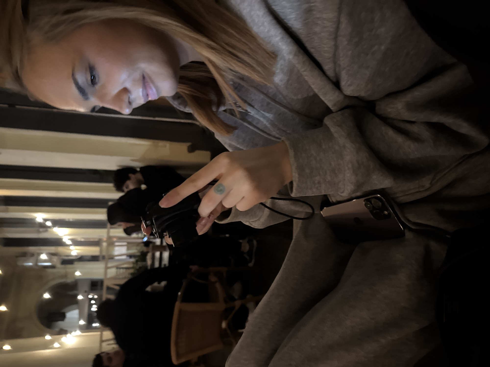 The image shows a medium shot of a woman sitting indoors, holding a black camera in her hands. She is looking down at the camera, her face partially obscured. She is wearing a grey hooded sweatshirt and grey sweatpants. A smartphone is tucked into her sweatpants. In the background, several other people are visible, as well as chairs and lights that suggest a dimly lit indoor setting. The environment appears to be a restaurant or cafe. There are no specific visual cues in the image to determine the location in the city of Warsaw, Poland.