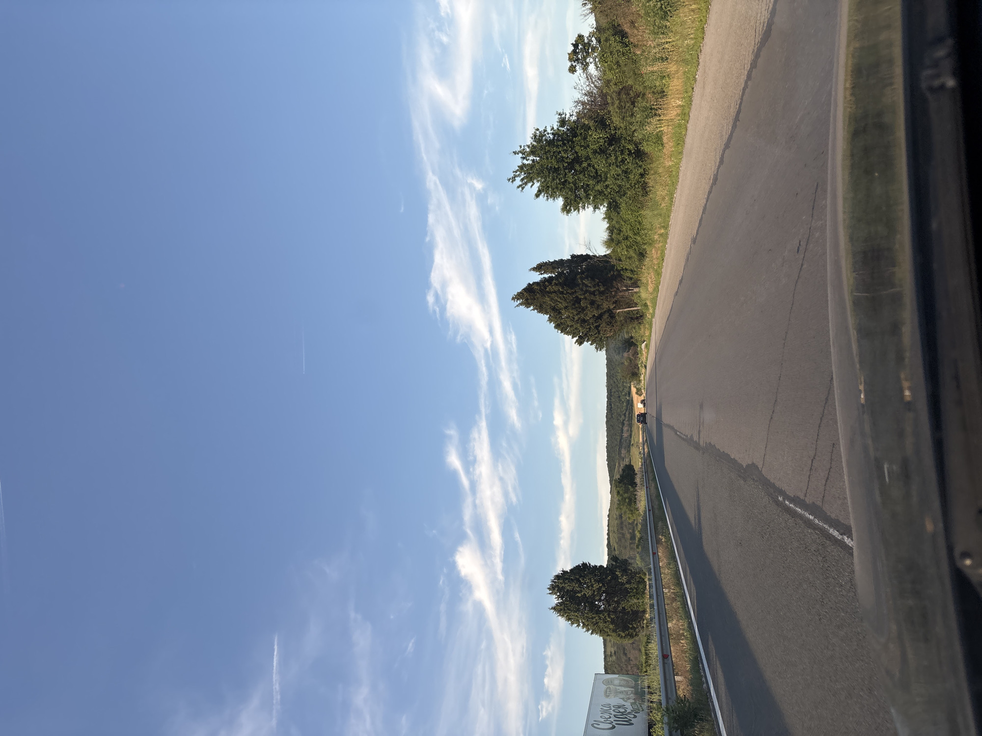 The image shows a view from a vehicle traveling on a paved road in Kipra, Bulgaria. The road stretches into the distance, with a slight curve to the right. On the left side of the road, a white guardrail separates the road from a grassy embankment and a billboard. Two cypress trees are prominent on the left, with a broader, leafy tree further down the embankment. The right side of the road is bordered by dense green vegetation and dry grass. In the distance, a small tunnel or underpass can be seen crossing the road, with vehicles approaching it. The sky is a clear blue with wispy white clouds, and two faint contrails are visible. The lower portion of the image shows the dashboard of the vehicle from which the photo was taken. The overall scene depicts a rural highway on a sunny day.