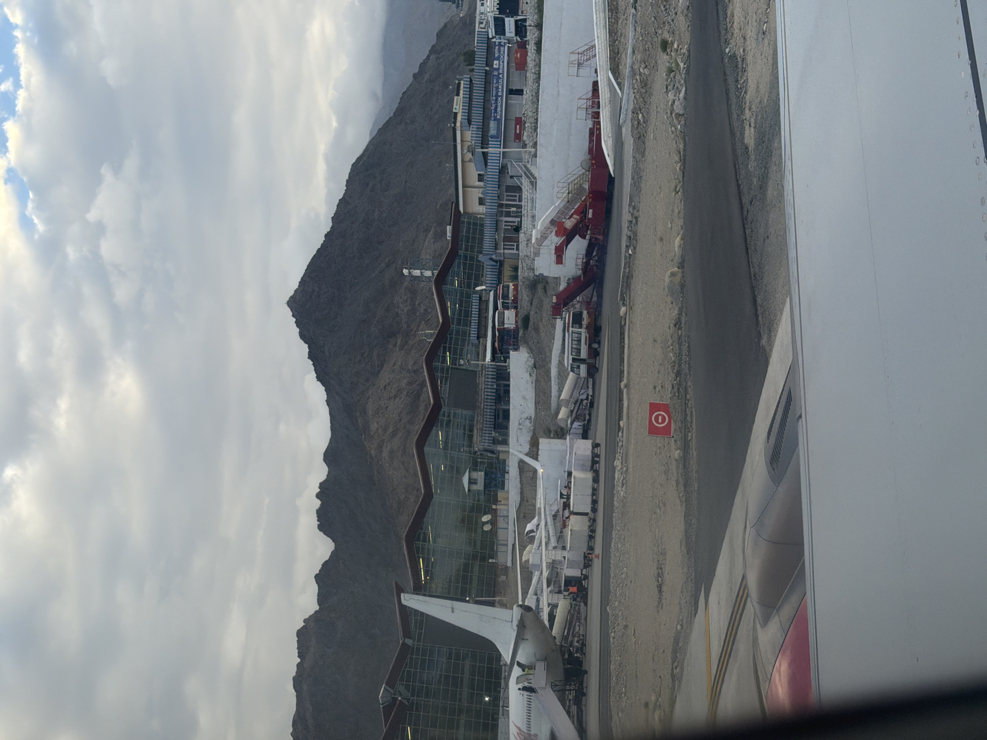 The image depicts Kushok Bakula Rimpochee Airport in Spituk, India, with a mountain range in the background under a cloudy sky. In the foreground, a portion of an airplane wing is visible. The airport tarmac shows an aircraft with its tail clearly visible, along with a boarding stairs and baggage carts. Several buses are also present on the tarmac. The airport building has a distinctive modern design with a glass facade. A red circular sign with a white horizontal bar, indicating a prohibition, is planted on the ground near the tarmac. The overall scene is of an active airport environment, with the rugged mountainous landscape serving as a dramatic backdrop.